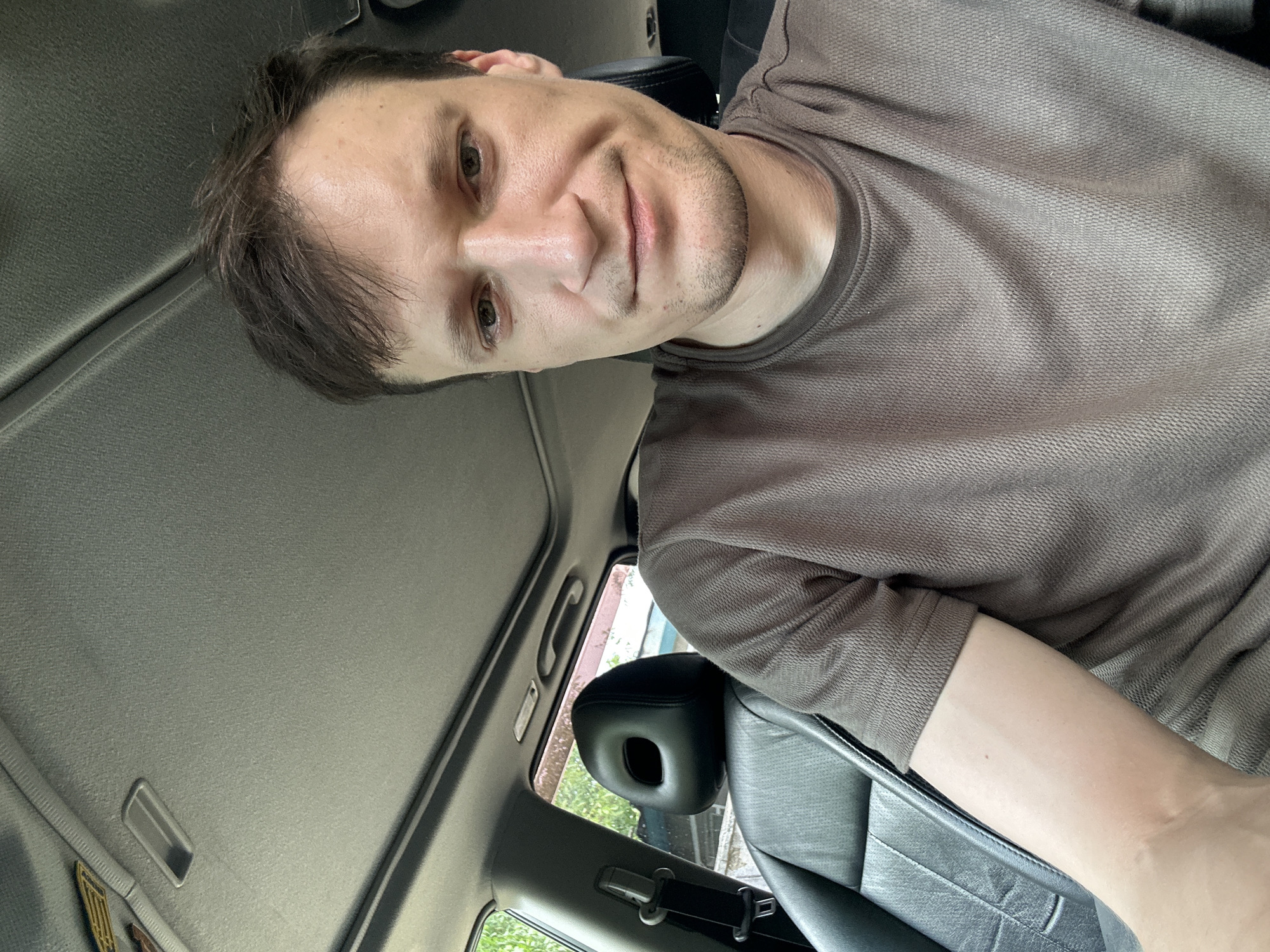 A man is seated in the driver's seat of a car, looking at the camera. The car's interior is visible, including a black leather seat and the ceiling with a sunroof. The man has short, dark brown hair and is wearing a brown t-shirt. The scene appears to be in an outdoor setting, possibly a street or parking lot in Makiv, Ukraine, suggested by the presence of trees and buildings visible through the car window. The lighting is natural, indicating it is daytime.