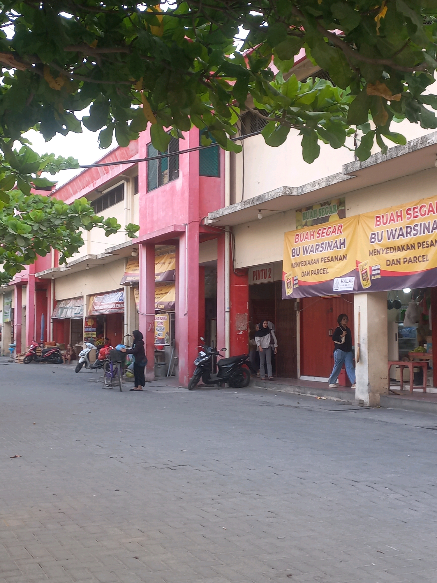 The image depicts a street scene in Baturetno, Indonesia, with a focus on a row of shops. The buildings are painted in bright colors, predominantly pink and cream. Large banners, likely advertising local businesses, are hung across the front of the shops, with text in Indonesian that appears to include "BUAH SEGAR BU WARSINAH."
Several people are visible in the scene. In the foreground, a woman in a black headscarf and dark clothing stands next to a bicycle, appearing to interact with the bicycle. Further down the street, a group of people are near an entrance labeled "PINTU 2," with some standing and others appearing to walk. Another woman in blue jeans and a dark top walks on the sidewalk.
Various objects are present, including motorcycles parked on the side of the street, a table and chairs outside one of the shops, and what appears to be produce or goods displayed within the shop entrances. The street itself is paved with interlocking bricks. Overhead, the branches of a tree with green leaves partially obscure the top of the buildings, suggesting an outdoor setting with vegetation. The overall impression is of a busy, local marketplace or commercial area.