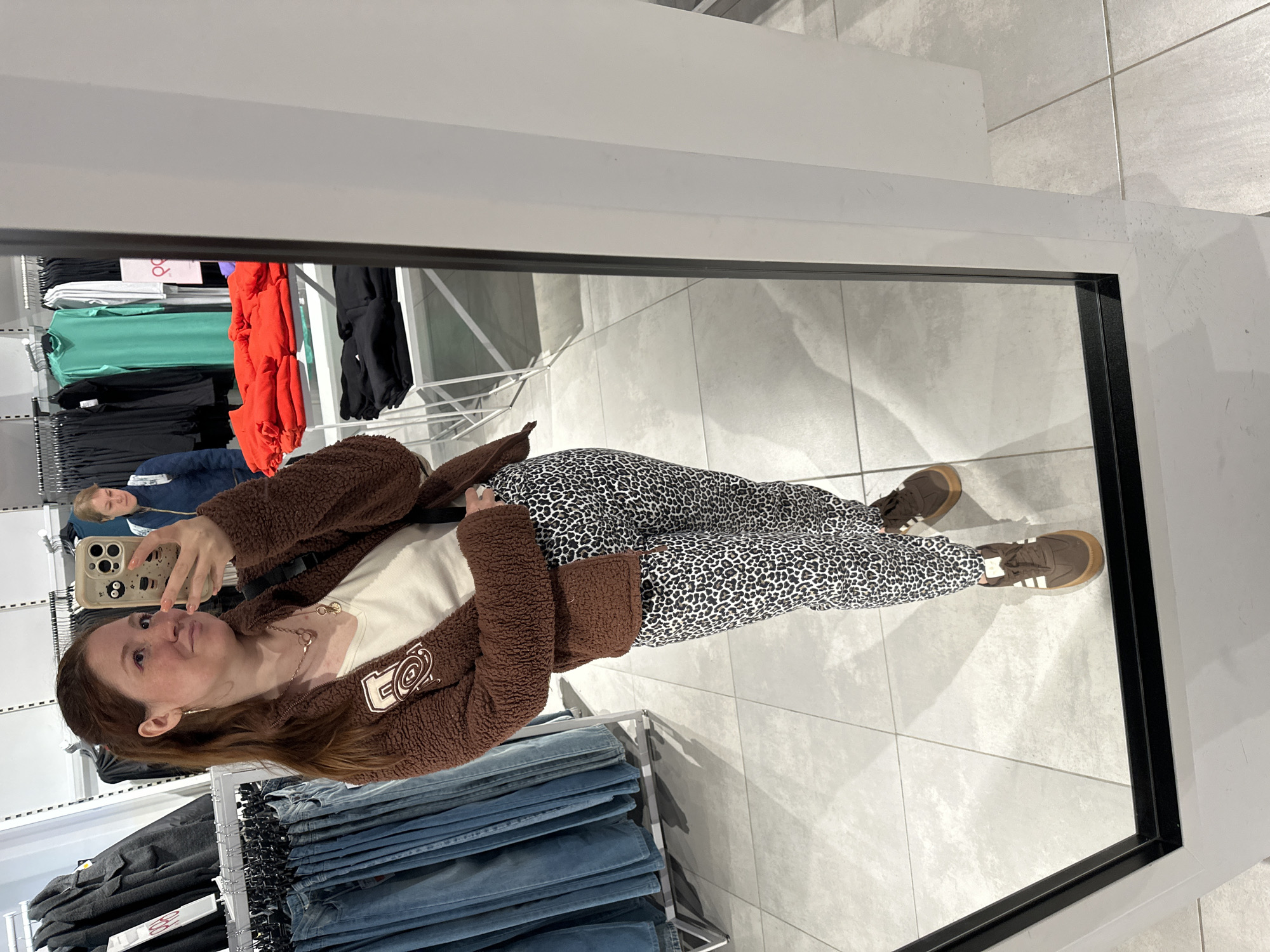 A person is reflected in a mirror in what appears to be a retail clothing store. The person is wearing a brown, fuzzy jacket, a cream-colored top, leopard-print pants, and brown sneakers with white stripes. The person is holding a smartphone in their left hand, presumably taking a selfie. The mirror reflects a view of clothing racks with various garments, including jeans, t-shirts in different colors (green, red, black, white), and other items. An individual in a blue shirt is also visible in the reflection. The floor is tiled in a light gray color. The location is unknown and cannot be identified as Nizhny Novgorod, Russia, from the visual information provided.