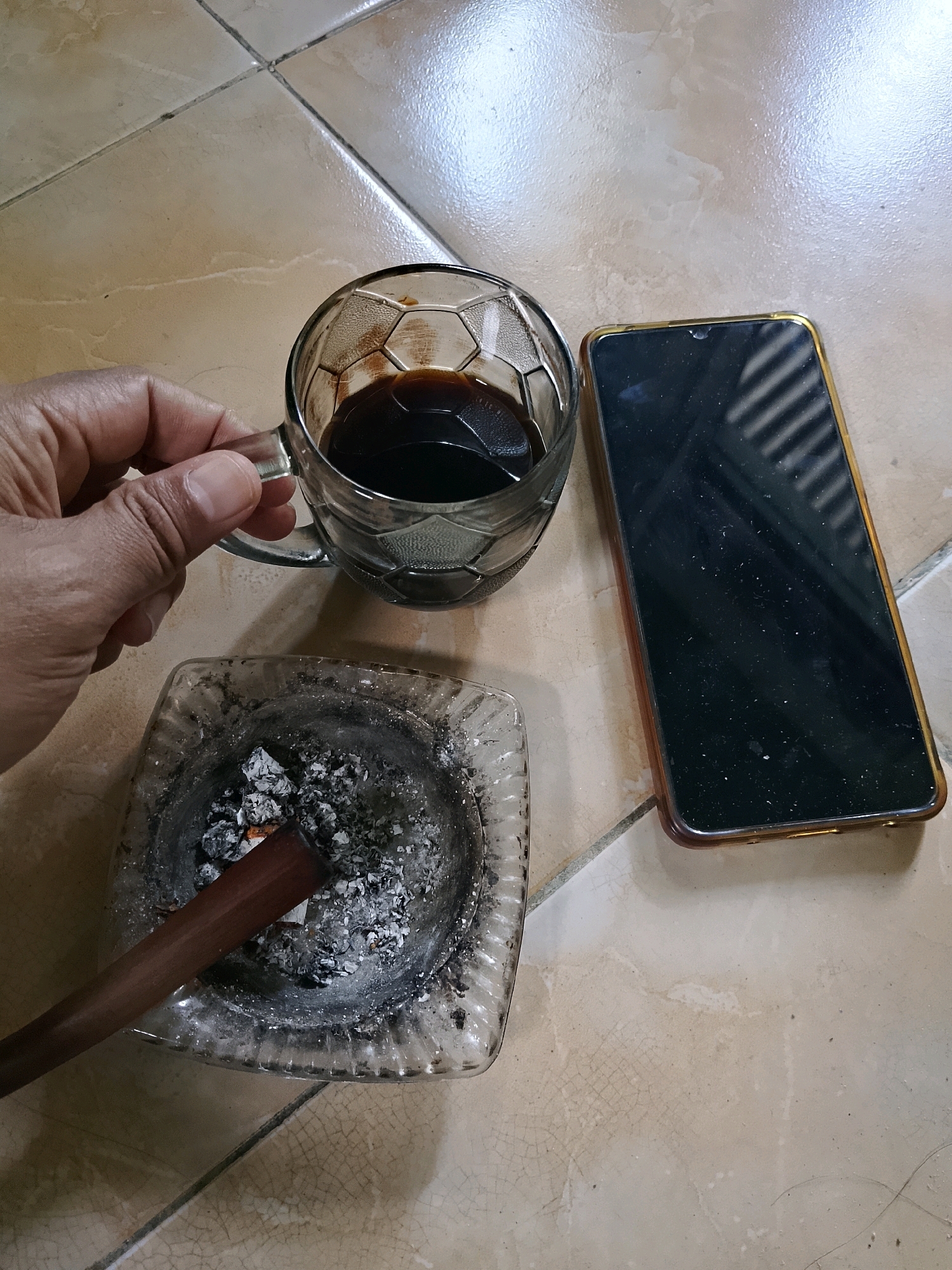 A top-down shot shows a person's hand holding a glass mug of dark liquid, likely coffee, with a hexagonal pattern etched into the glass. Beside the mug is a clear glass ashtray containing cigarette ash and a half-smoked cigar. To the right of the ashtray and mug is a smartphone with a black screen and a gold-colored frame. The objects are arranged on a tiled floor with a light beige color and visible grout lines. The lighting suggests an indoor environment. The scene appears to be a casual break, possibly in a home or a relaxed setting in Jember, Indonesia.