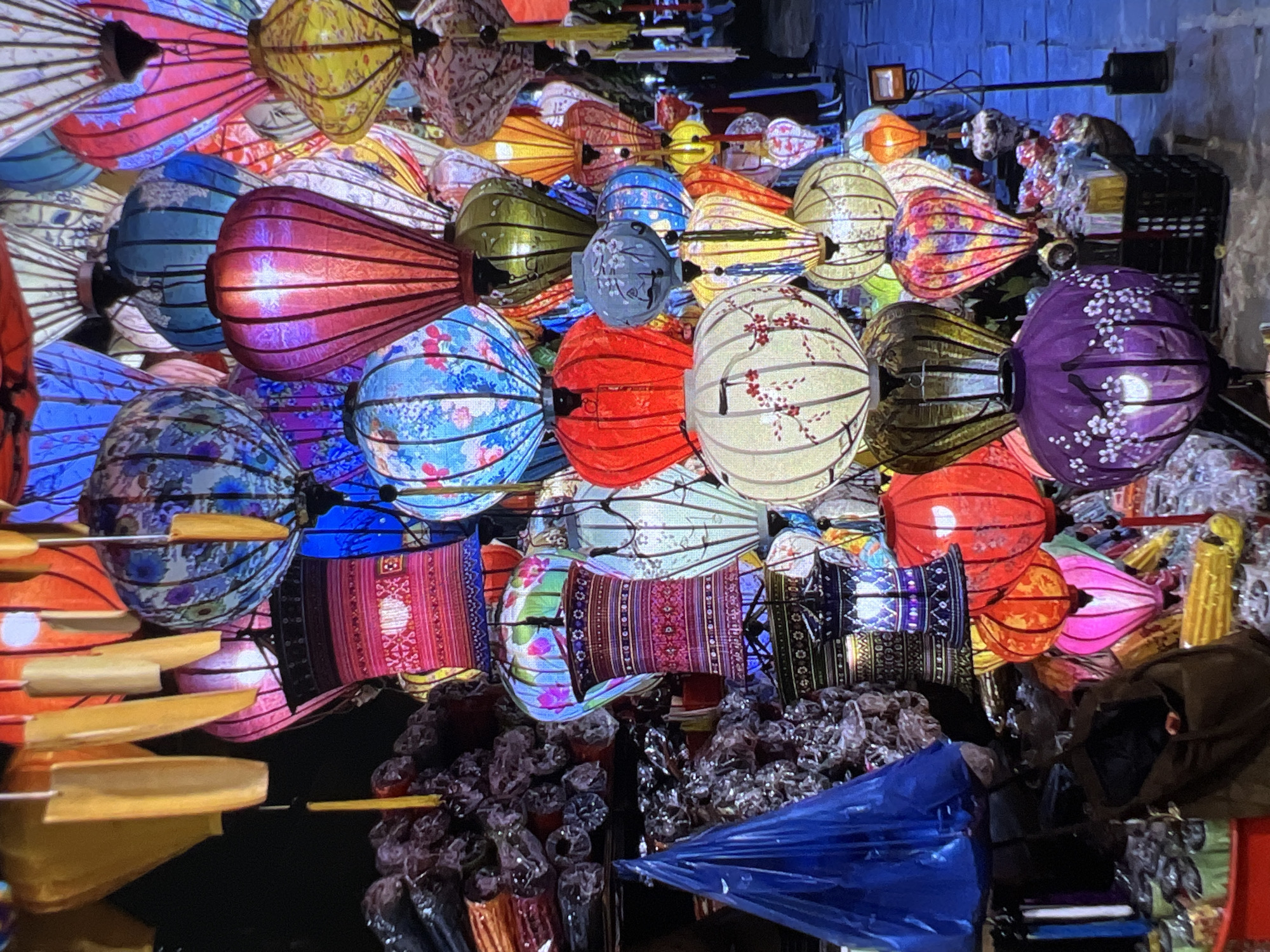The image displays a vibrant and densely packed collection of illuminated, colorful lanterns, likely at a market or festival. The lanterns are of various shapes, including round, elongated, cylindrical, and teardrop, and come in a rich palette of reds, blues, purples, yellows, greens, and oranges. Many feature intricate patterns, such as floral designs, geometric motifs, and delicate line work, often appearing to be made of silk or paper with visible internal structures.
The intense glow from within the lanterns suggests they are illuminated in a low-light setting, such as nighttime or a dimly lit interior space, highlighting their luminescence. They are arranged closely together, creating a tapestry of light and color. In the lower sections of the image, bundles of plastic-wrapped items, possibly smaller lanterns or other merchandise, are visible, alongside dark bags or crates and a bright blue plastic bag. Some bamboo-like sticks or handles are also present, which might be for carrying or displaying the lanterns.
While no people are visible, the scene implies a bustling atmosphere of commerce or celebration. The style and craftsmanship of the lanterns strongly suggest a cultural context from an East or Southeast Asian country, where such traditional lanterns are commonly used for decoration, particularly during festivals. The overall mood is one of festivity, cultural richness, and visual spectacle. No discernible text is visible in the image.