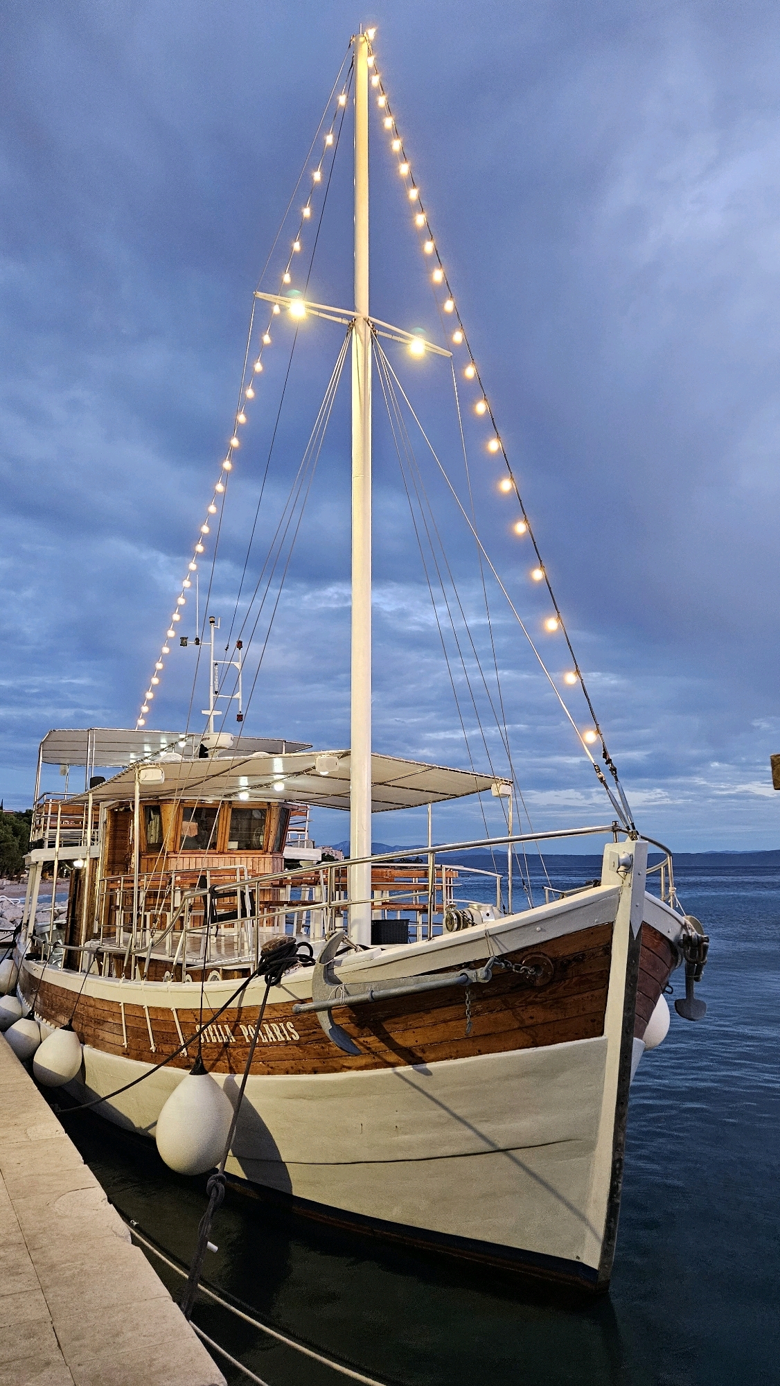 A wooden boat with white accents is docked by a stone walkway. The boat is illuminated by a string of yellow lights that run along the mast and rigging. The vessel has a covered upper deck with seating and a wooden cabin structure. The boat is tied to the dock with ropes and fenders. The water appears dark and calm, and the sky is overcast with dramatic clouds. The scene suggests a coastal location, possibly in Općina Tučepi, Croatia, given the typical Mediterranean aesthetic.