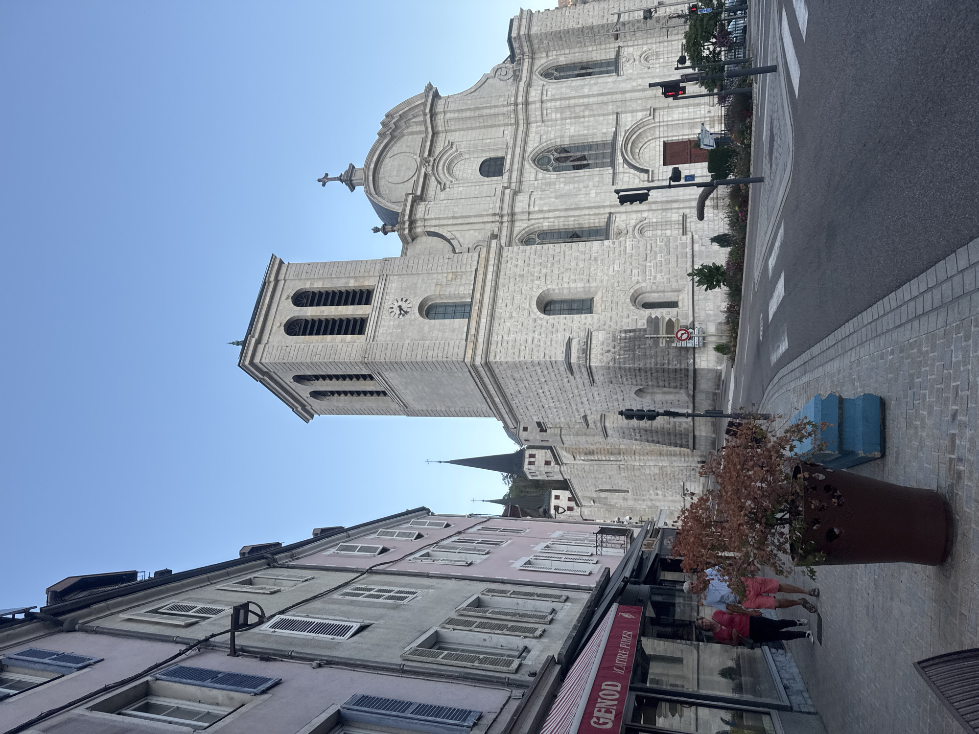This image captures a street scene in the city of Saint-Claude, France, featuring the Saint-Pierre Cathedral as the dominant structure. In the foreground, a paved sidewalk runs alongside a building with a pink facade and windows with closed shutters. A business with a red and white striped awning, labeled "GENOD MAITRE PIMER," is visible. Two individuals, a man and a woman, are walking on the sidewalk away from the camera. The man is wearing a light blue shirt and pink shorts, while the woman is wearing a red shirt and black pants. A large brown planter with a small tree containing reddish-brown leaves is positioned on the sidewalk, along with a blue concrete barrier.
The street itself is asphalt, with a pedestrian crossing marked by white stripes. Traffic lights are visible at the intersection. To the right of the sidewalk and street, the imposing stone facade of the Saint-Pierre Cathedral rises. The cathedral has a tall bell tower with arched openings and a clock. Further back and to the left, other spired structures, possibly part of the cathedral complex or other nearby buildings, are visible against a clear blue sky. The overall lighting suggests it is late afternoon or early evening.
