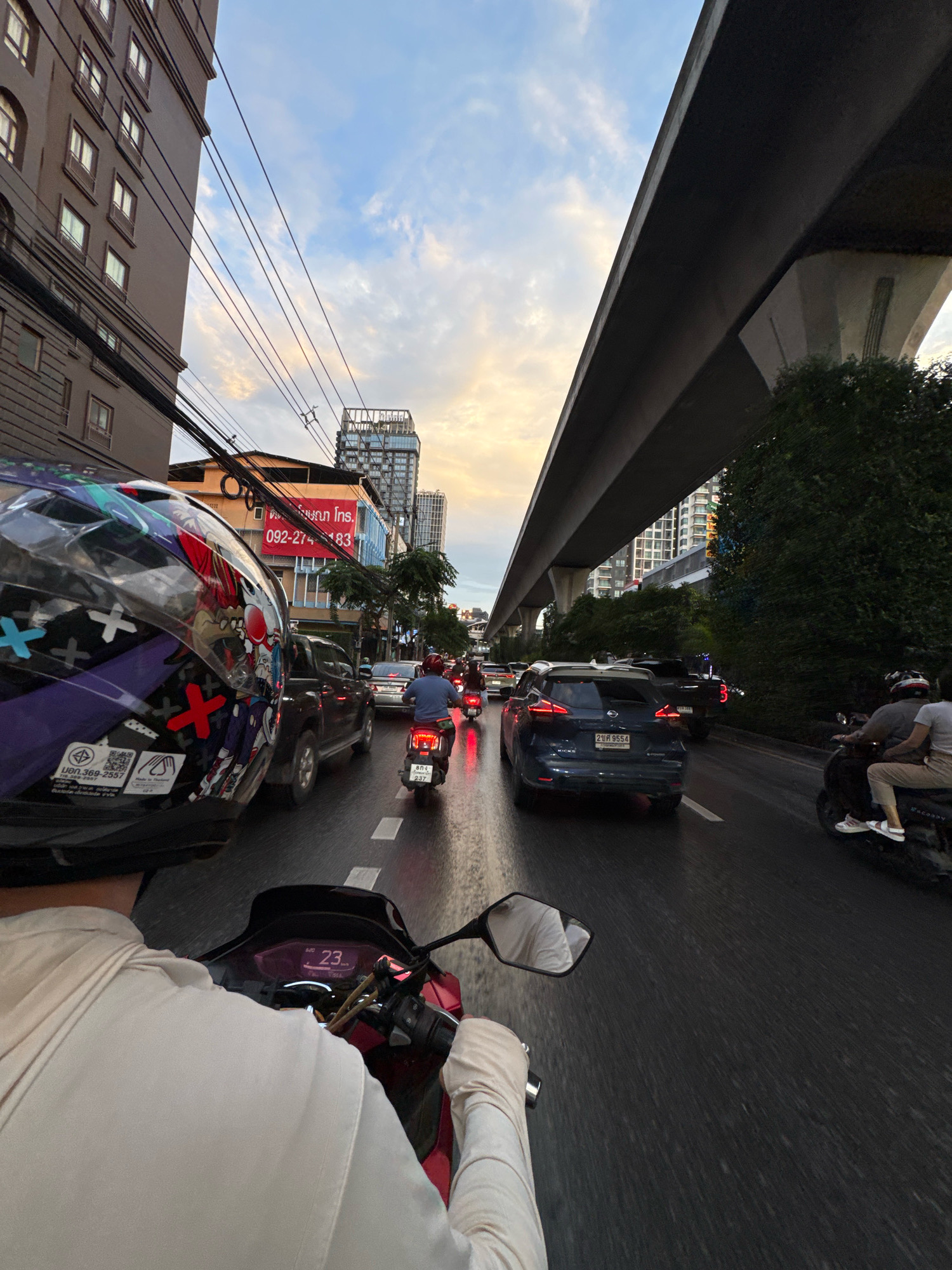 The image depicts a first-person perspective of someone riding a red motorcycle through congested traffic in Bangkok, Thailand, during the late afternoon or early evening. The rider is wearing a white long-sleeved shirt and a helmet decorated with a colorful graphic pattern. The motorcycle's digital display shows "23" kilometers per hour.
The scene is characterized by a multi-lane road with several vehicles, including cars and other motorcycles, all moving slowly in traffic. On the left side of the road, there are tall, multi-story buildings with numerous windows. A prominent elevated highway structure dominates the upper right portion of the image, casting a shadow over the road. Trees and greenery are visible beneath the highway.
A large red banner with Thai text and a phone number, "092-274-3183," is visible on a building in the background, suggesting a commercial area. The sky is a mix of blue with some scattered clouds, illuminated by the setting or rising sun, creating a warm, diffused light. The road surface appears wet, indicating recent rain or ongoing light precipitation. The overall impression is one of urban traffic, typical of Bangkok's busy thoroughfares.