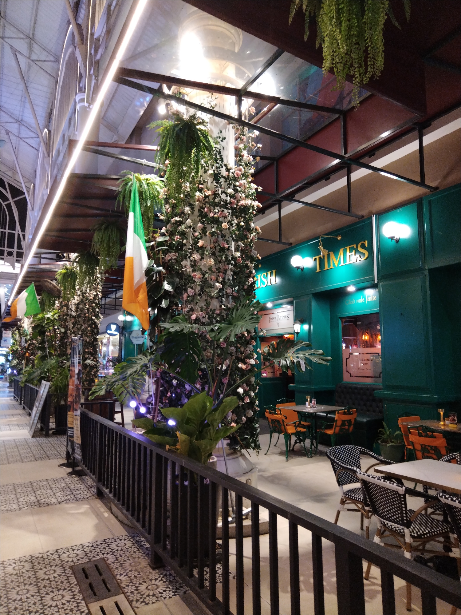 The image shows the exterior of an establishment in Patong, Thailand, identified by signage as "Irish Times". The building is painted a distinctive dark green. Two Irish flags are visible, one prominently displayed on a decorative floral column in the foreground, and another smaller one further back. The facade features large windows revealing an interior with seating and lighting. Outdoor seating with tables and chairs, some orange and others with black and white patterned seats, is arranged on a tiled patio area. The area is decorated with numerous plants, including hanging ferns and large potted plants, creating a lush, green atmosphere. Overhead, a covered walkway with modern lighting and a metal structure can be seen. The overall scene depicts a restaurant or pub with a clear Irish theme, situated in an outdoor or semi-outdoor setting.