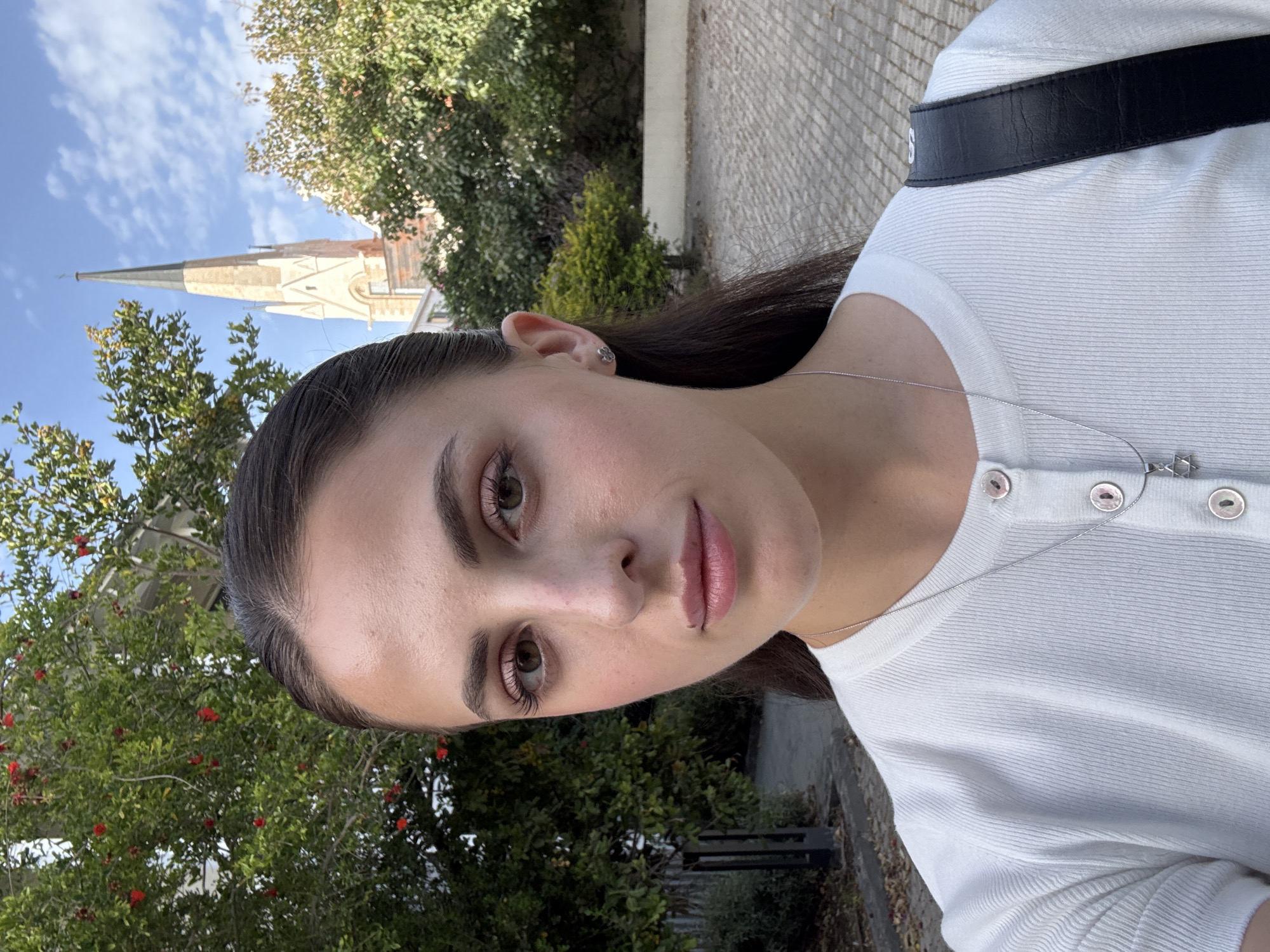 A close-up, slightly rotated selfie of a young, fair-skinned woman with brown hair and eyes. She is wearing a white, ribbed, button-down shirt, and a silver necklace with a Star of David pendant. She has a nose piercing on the left side. Behind her, there are trees with red berries and a paved street. In the upper left corner of the image, the top of a church steeple is visible against a blue sky. Based on the architecture of the church, the vegetation, and the urban landscape, it appears that this photo may have been taken in the city of Tel Aviv, Israel.