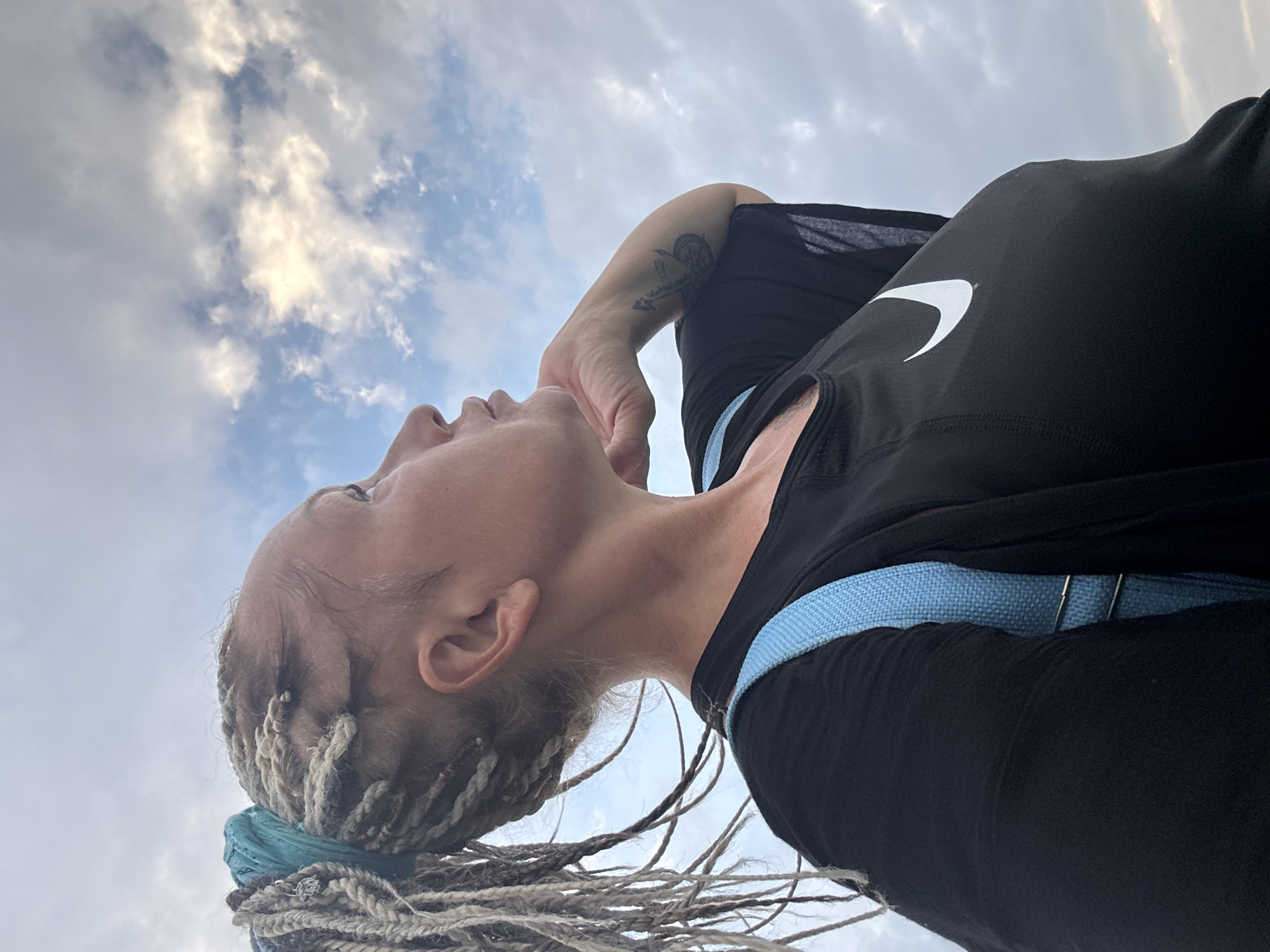 A woman with blonde dreadlocks pulled back with a turquoise fabric stands outdoors, looking up at a cloudy sky. She wears a black t-shirt with a white Nike logo and a light blue strap over her shoulder. Her right arm is bent, and her hand rests on her chin. A tattoo is visible on her forearm. The background is a sky filled with white and gray clouds. The scene is captured from a low angle, looking upwards.
