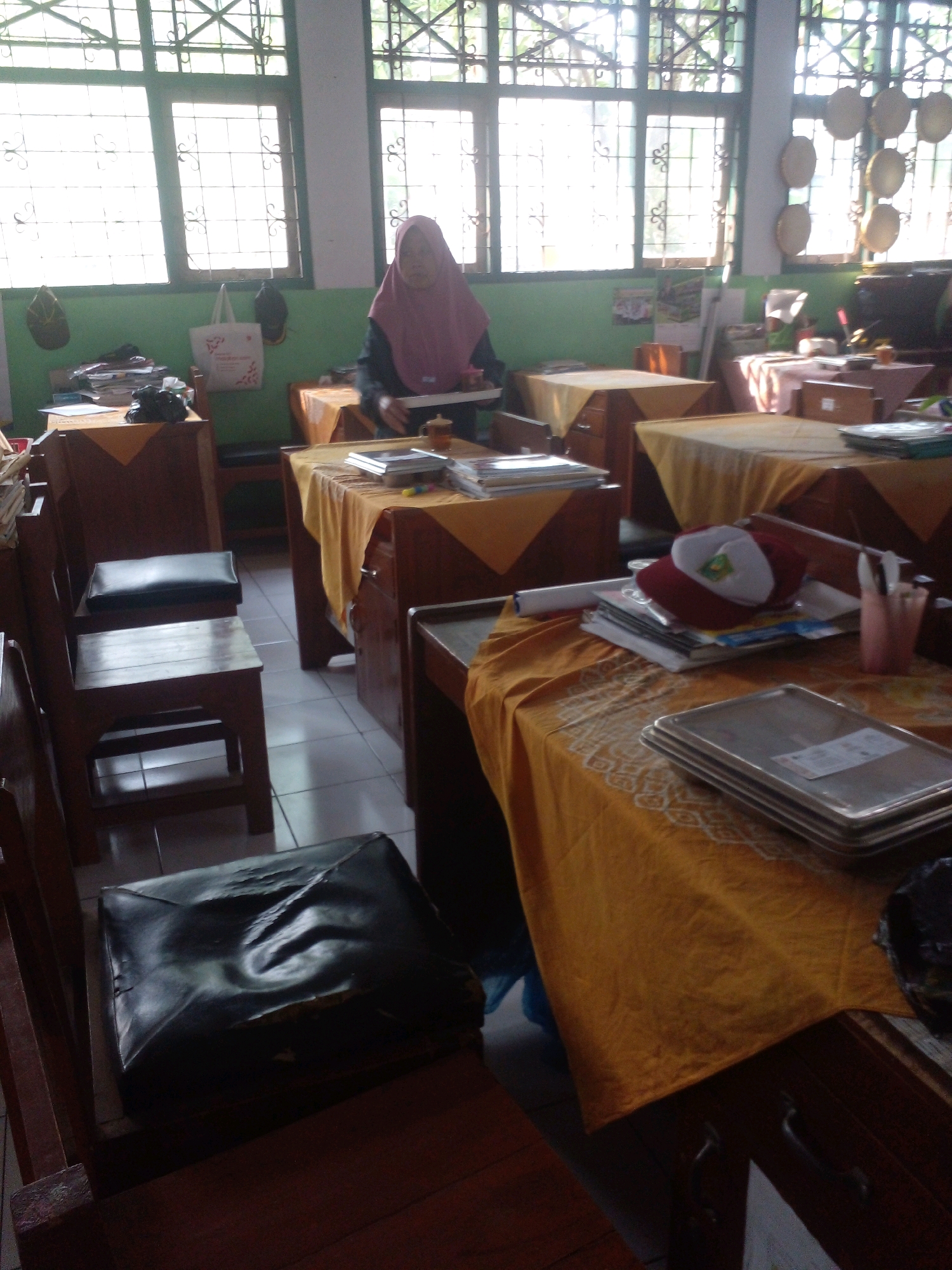In a classroom in Banyuputih, Indonesia, a student wearing a pink hijab sits at a wooden desk. The classroom has large windows with ornate metal grilles, and the walls are painted green. The desks are covered with yellow tablecloths. On one desk, there are stacks of books, a red and white hat, and a pink cup with pens. On another desk, there are trays and books. The floor is tiled.