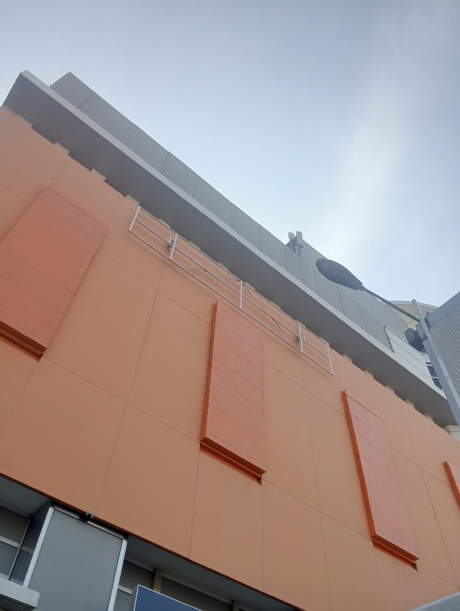 This is a low-angle, medium shot of the exterior of a modern building under a cloudy sky. The building's facade is primarily a pale orange color, divided by faint grid lines. Several rectangular panels, also orange but with a textured appearance, are vertically mounted on the facade. A white metal railing or scaffolding structure is visible across the upper part of the orange section. Above the orange section is a gray section of the building with a corrugated texture, and a black street lamp is mounted on the upper level, extending over the facade. At the very top, a sliver of a lighter gray building is visible. The bottom of the image shows a portion of the building's lower level with large, rectangular windows or panels that appear to be gray and silver. The sky is overcast and pale. The architecture suggests it is a commercial building, likely a shopping mall or office complex. Based on visual cues, this building is likely located in the city of Surabaya, Indonesia.