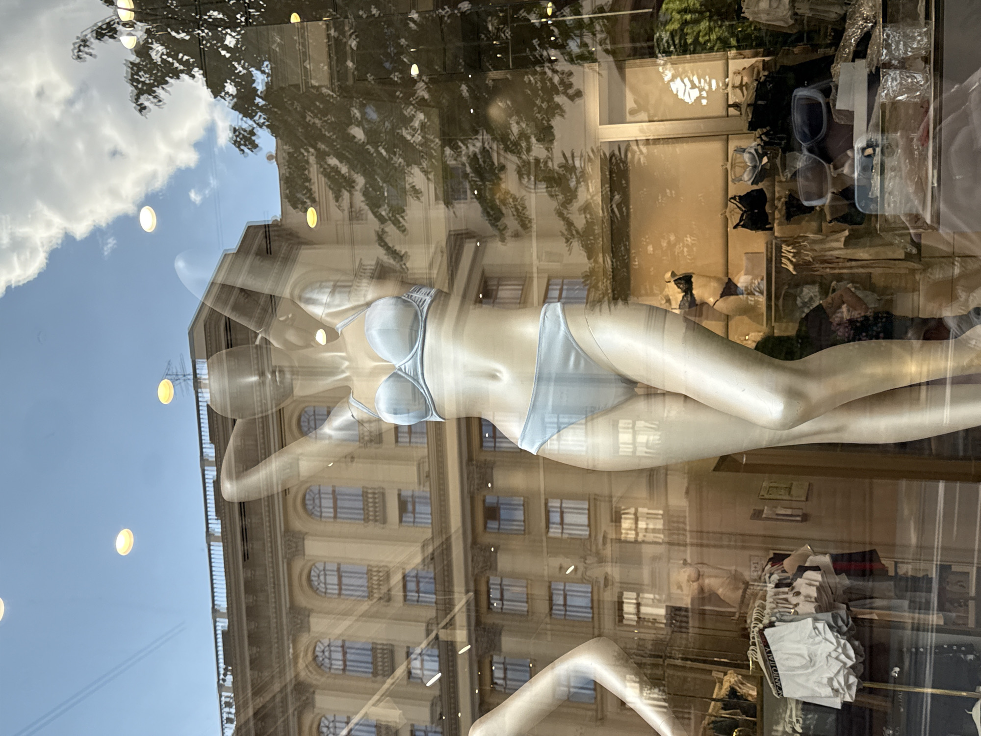The image shows a reflection in a storefront window, depicting a mannequin wearing light blue lingerie. Behind the mannequin, a building with classical architecture, featuring arched windows and ornate detailing, is visible. The sky, with scattered clouds and several circular lights, is also reflected in the glass. In the foreground and to the left of the mannequin, other store merchandise, including men's underwear and bras, are displayed on racks. The scene appears to be a retail display in a city. Given the architectural style and the presence of Cyrillic text on some of the displayed items, this location is likely Moscow, Russia.