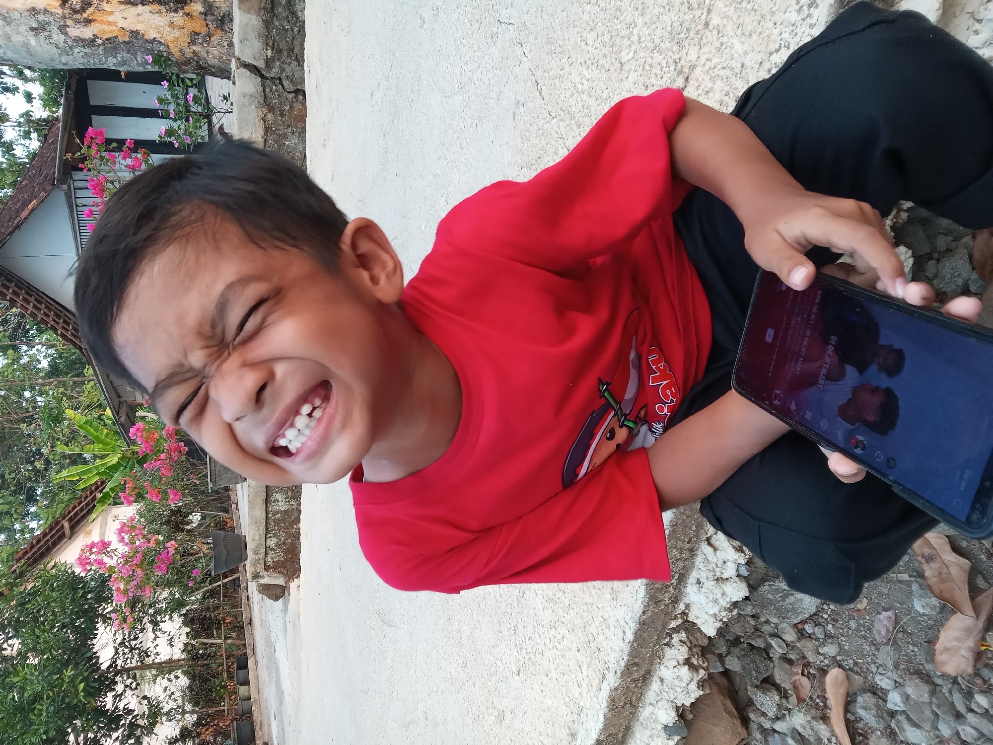A young Indonesian boy, wearing a red t-shirt and black pants, sits on a concrete surface outdoors. He is holding a smartphone in his right hand and making a playful face, scrunching his eyes and baring his teeth. The background shows a modest house with a white and brown facade, surrounded by greenery and flowering plants. The scene appears to be in a rural or suburban area, possibly in Kedunggudel, Indonesia, given the tropical vegetation and architectural style. The ground is a mix of concrete, rocks, and dry leaves.