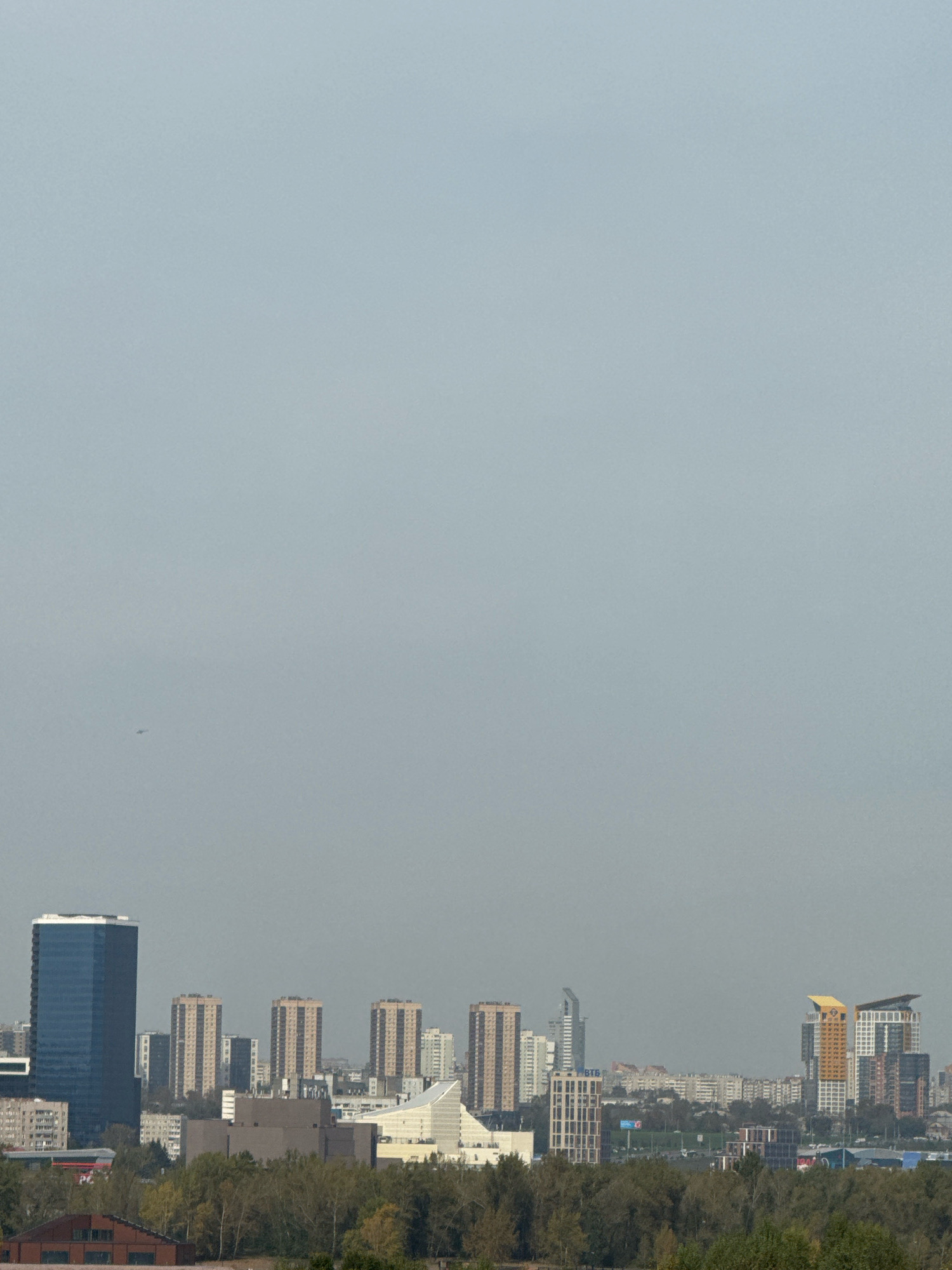 The image depicts a panoramic view of a city skyline under a hazy, overcast sky. In the foreground, a dense line of trees with autumn foliage lines the bottom of the frame, with a few low-rise buildings, including a red-brick structure with a gabled roof, visible amongst the trees. In the mid-ground, a collection of modern buildings, including several high-rise apartment blocks and office buildings, forms the cityscape. A distinctive dark blue glass skyscraper stands prominently on the left side of the frame. Further to the right, a cluster of beige apartment buildings with visible balconies are arranged in a row, and beyond them, more buildings of varying heights and architectural styles extend into the distance. Notably, on the far right, two brightly colored, modern skyscrapers, one yellow-topped and another with a white facade, punctuate the skyline. In the far distance, the city extends further, with numerous buildings and what appears to be a bridge or elevated structure visible. The overall impression is one of a sprawling urban environment. This scene is consistent with a view of the Pokrovsky district in Krasnoyarsk, Russia, looking towards the northeast.
