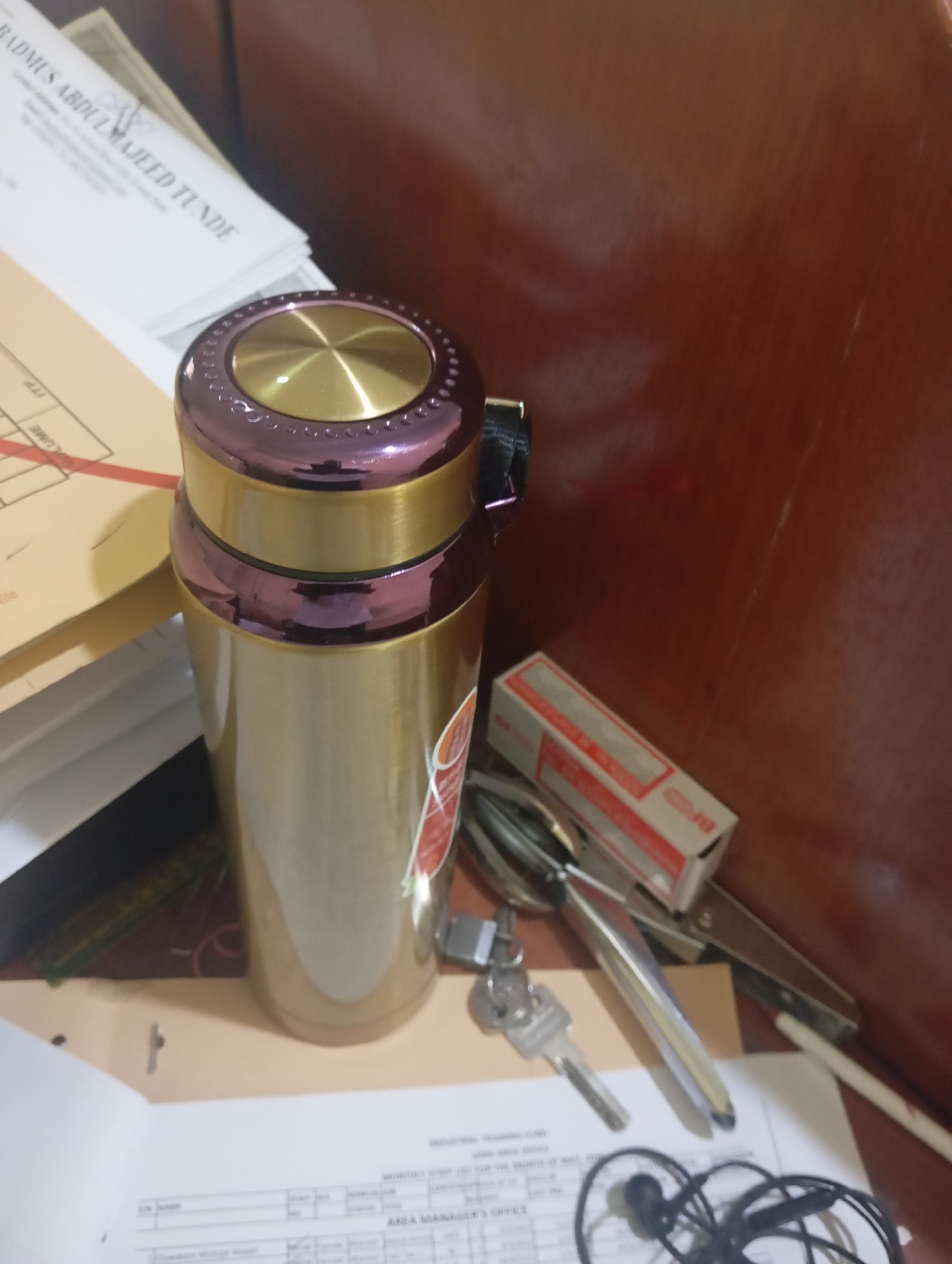 A high-angle, close-up shot focuses on a gold and purple thermos bottle with a metallic lid. The thermos is positioned on a wooden desk cluttered with various office supplies. To the left of the thermos, stacks of papers are visible, one with legible text reading "BADMUS ABDULMAJEED TUNDE". To the right of the thermos, a small red and white box, what appears to be a stapler, a pair of tweezers, and a key are scattered on the desk. In the foreground, the corner of a document with tabular data is visible, and a pair of earbuds with tangled wires rests on the desk. The background is dominated by the dark, warm tones of a wooden surface. The lighting appears to be artificial, casting soft shadows. This scene is consistent with an office environment, possibly in Jos, Nigeria, given the subject's name printed on the document, but no specific landmarks or indicators of Jos are visible within the frame.