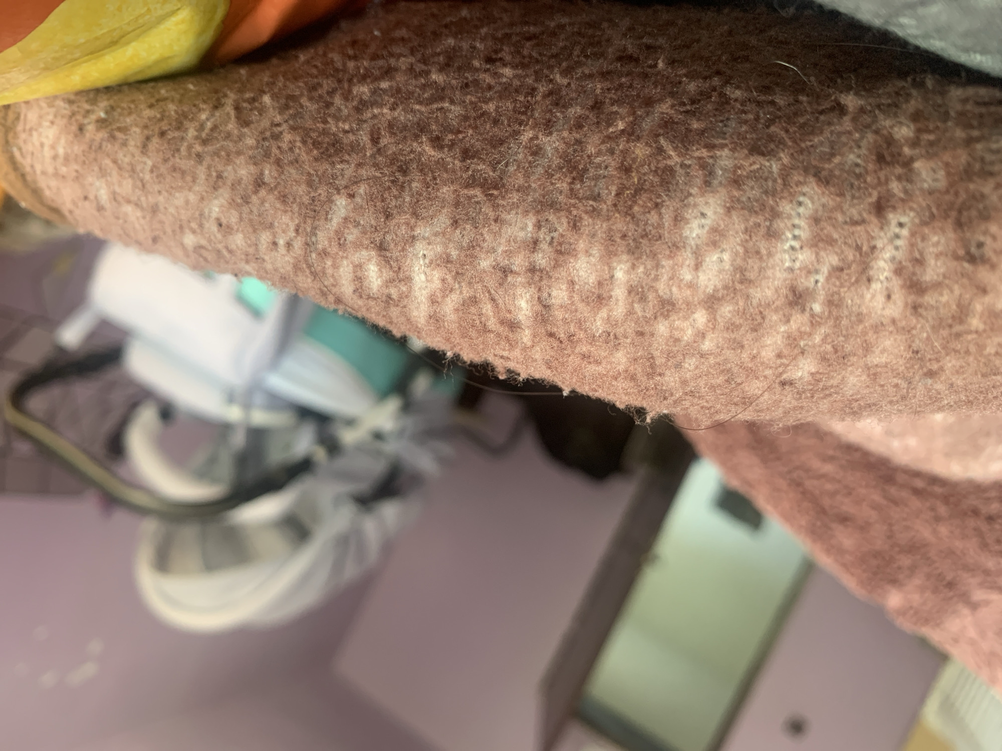 The image shows a close-up view of a brown knitted fabric, possibly a blanket or sweater, with visible individual fibers and a subtle pattern of lighter colored threads woven in. In the background, out of focus, a room is visible with purple walls. A doorway with a light-colored door is seen on the left, and on the right, a baby stroller or carrier with a white canopy is present. The scene appears to be indoors, in a domestic setting. No specific location within Krushev Dol, Bulgaria, can be determined from the image.