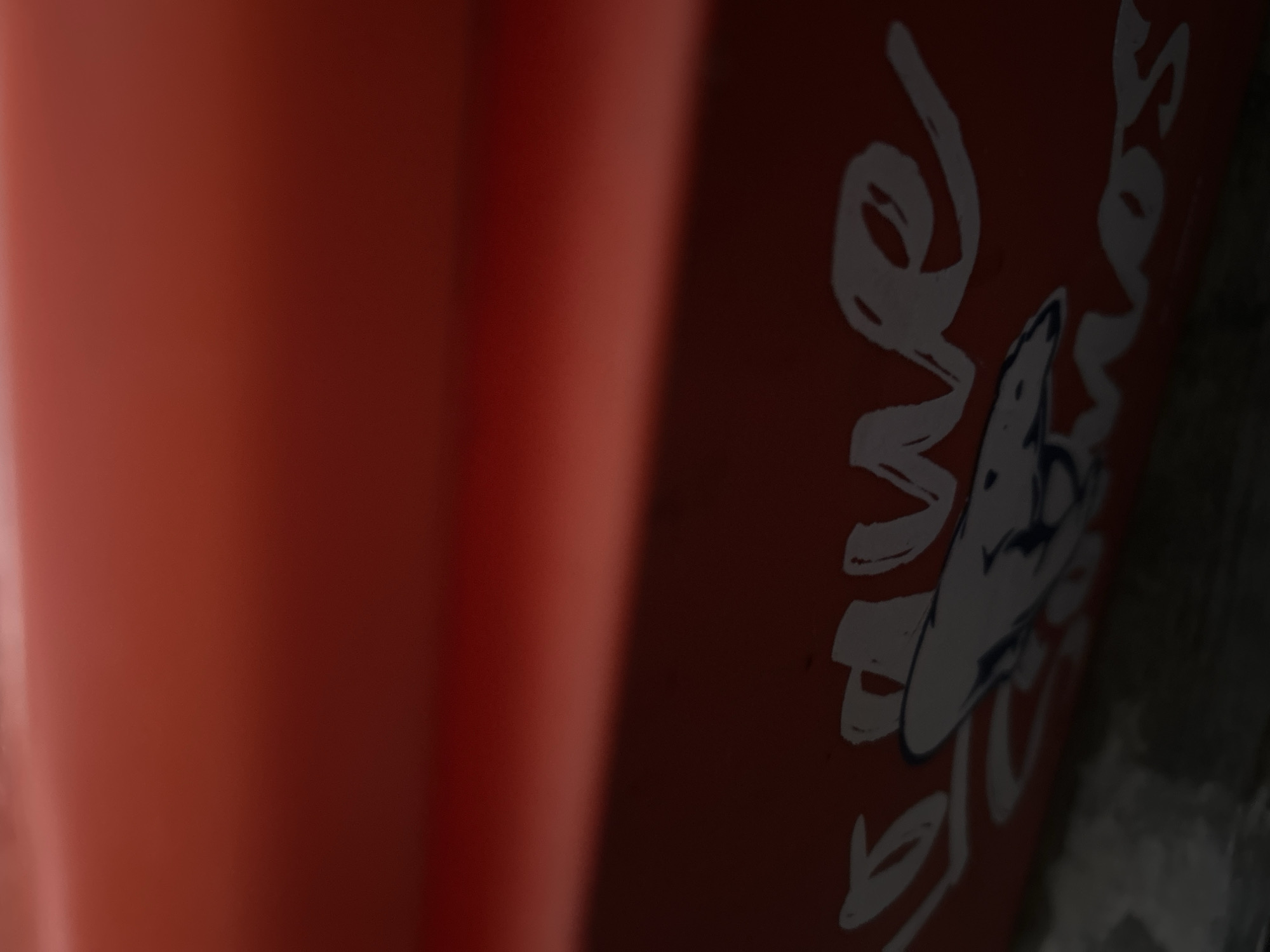 The image displays a close-up, angled view of a red surface with white lettering and a graphic. The lettering appears to be a stylized script, with portions of the words "Droguería" and "Cosmos" visible. A graphic element resembling a stylized animal or bird, possibly a shark, is depicted in the center. The surface has a glossy finish, reflecting ambient light. The bottom of the image is darker, suggesting a shadowed area or a change in surface texture. The context provided indicates this scene is located in Santa Ana, Venezuela.