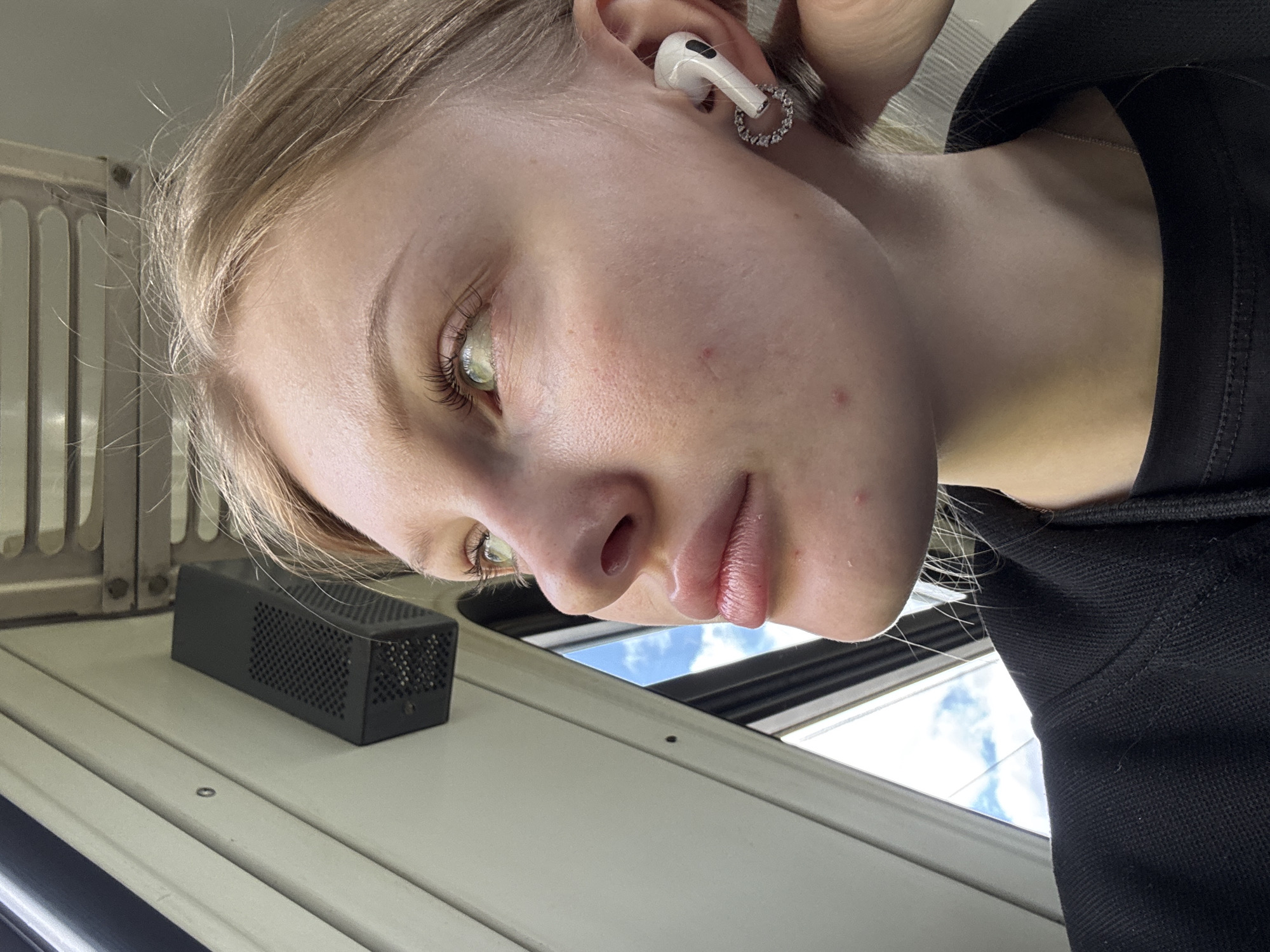 A close-up, slightly angled photograph depicts a young woman with fair skin, light blonde hair, and light-colored eyes, possibly green. She is wearing a white earbud in her left ear, adorned with a circular, possibly jeweled, earring attachment. She is wearing a black shirt, and a delicate silver chain is visible around her neck.
The environment appears to be an interior space with white or light-colored metal panels. A small, rectangular black object with a perforated surface rests on the panel to her left. Through a window, a glimpse of a blue sky with white clouds is visible. The image doesn't provide any additional information that would definitively place this scene in Mysy, Russia.