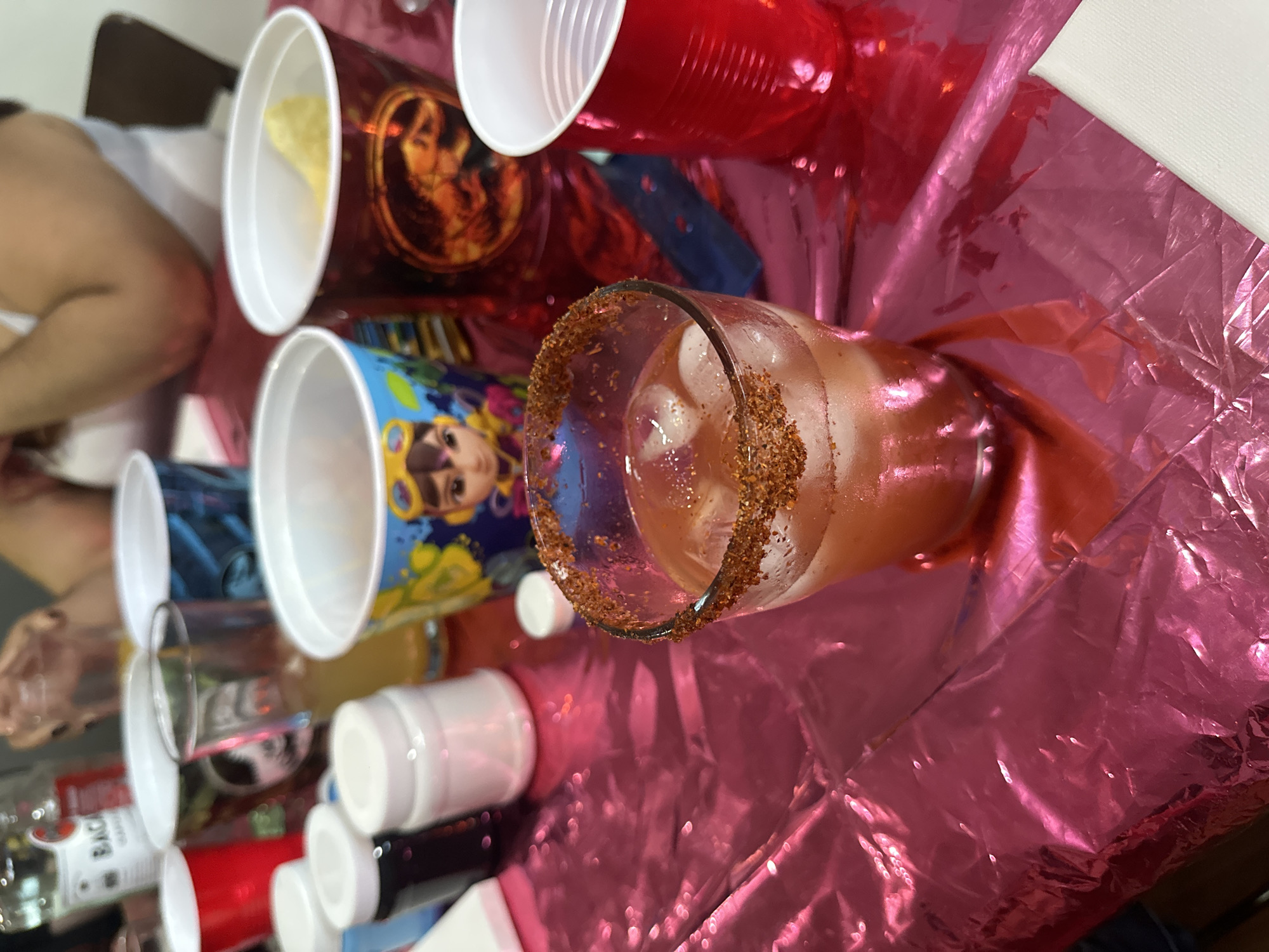 The image shows a gathering indoors with a vibrant pink tablecloth as a backdrop. Several plastic cups, both red and decorated with images, are scattered across the table. A glass filled with ice and a reddish liquid, garnished with a dark rim of what appears to be chili powder or seasoning, dominates the center of the frame. Assorted bottles are visible, including one that appears to be Bacardi rum. The setting gives the impression of a party or casual social event. There is no location information provided and therefore Unknown, Unknown is irrelevant.
