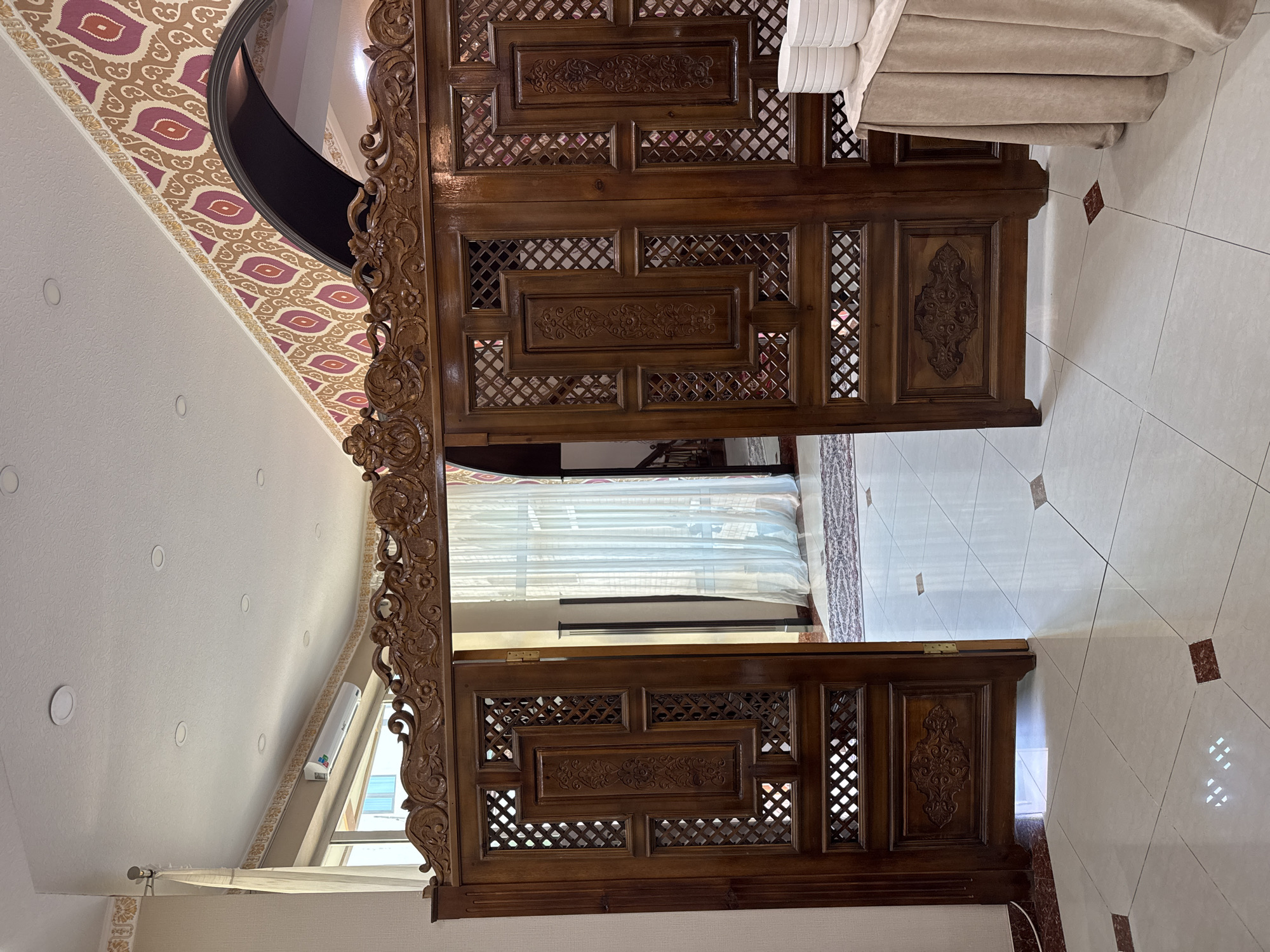 The image shows a wooden doorway or screen in what appears to be a room in Samarkand City, Uzbekistan. The doorway features intricate carvings and latticework. The floor is tiled, and there is patterned wallpaper visible near the ceiling. Through the doorway, a window with sheer curtains is visible.