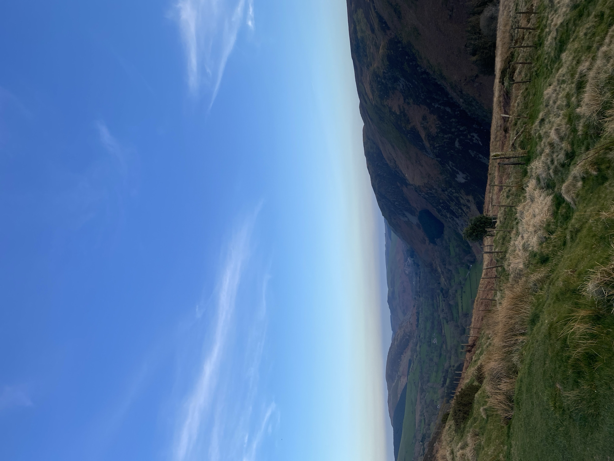 The image presents a landscape scene, likely in the vicinity of Llangynog, United Kingdom. It features a verdant hillside descending sharply into a valley. The grassy terrain is punctuated by a rustic wooden fence running along the slope. A prominent mountain dominates the upper right portion of the image, its steep face showing variations in vegetation. Below the mountain, a valley unfolds with a blend of green fields and forested areas. The sky transitions from a clear blue at the top left to a lighter hue near the horizon, with sparse, thin clouds.