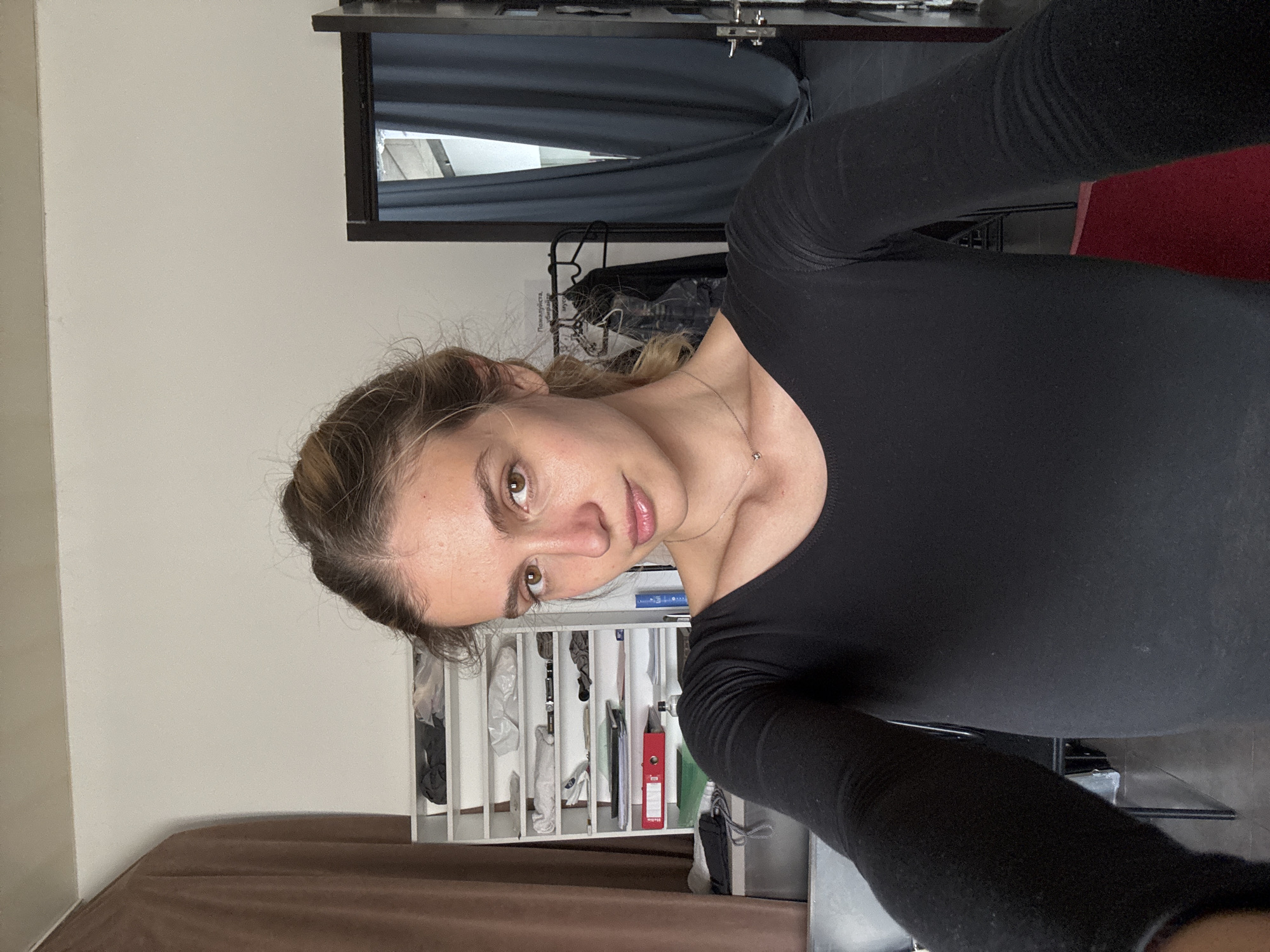 A woman is captured in a half-body selfie, oriented sideways in the frame. She has light brown hair pulled back with some loose strands, and brown eyes. She wears a plain black long-sleeved top and a delicate silver necklace with a small pendant. Her expression is neutral to slightly smiling.
The background reveals an indoor setting. To the upper left, a mirror reflects a dark curtain or fabric and a brightly lit area, possibly a window. Below the mirror, a white shelving unit is visible, filled with various items like clothes, bags, and what appears to be a red folder or binder. A black clothing rack or stand is situated behind her right shoulder. A piece of dark brown furniture or a curtain is at the bottom left, and a red object, possibly a chair or cushion, is partially visible on the right side. On a piece of paper hanging on the clothing rack, the Cyrillic word "Пожалуйста," (meaning "Please,") is discernible, along with other partially obscured text. The overall impression is of a casual indoor environment, possibly a personal room or a functional space like a changing room or studio.