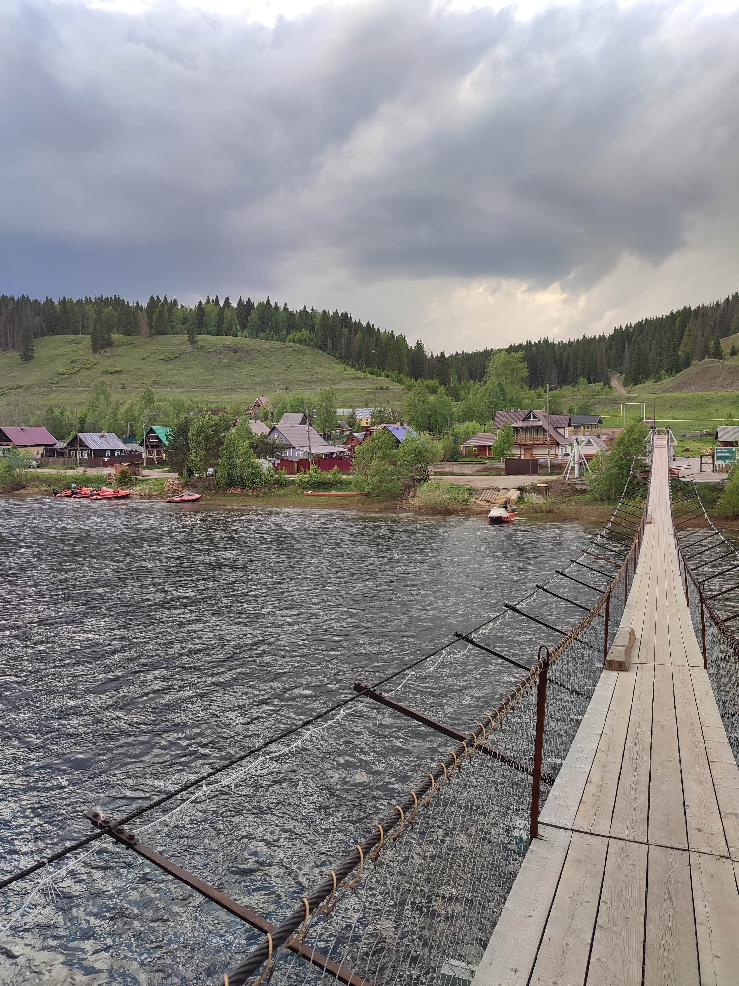 The image depicts a suspension bridge crossing a body of water, likely a river, in Усьва, Russia. The bridge is constructed of wood planks for the walkway and metal cables for support and railings. The water appears choppy, reflecting the overcast sky. In the background, there is a village with houses and buildings, surrounded by green hills and forest. Several boats are docked along the riverbank. The sky is filled with gray clouds, suggesting an overcast or possibly rainy day. The scene appears to be a rural or semi-rural setting. There are no people visible in the immediate scene.