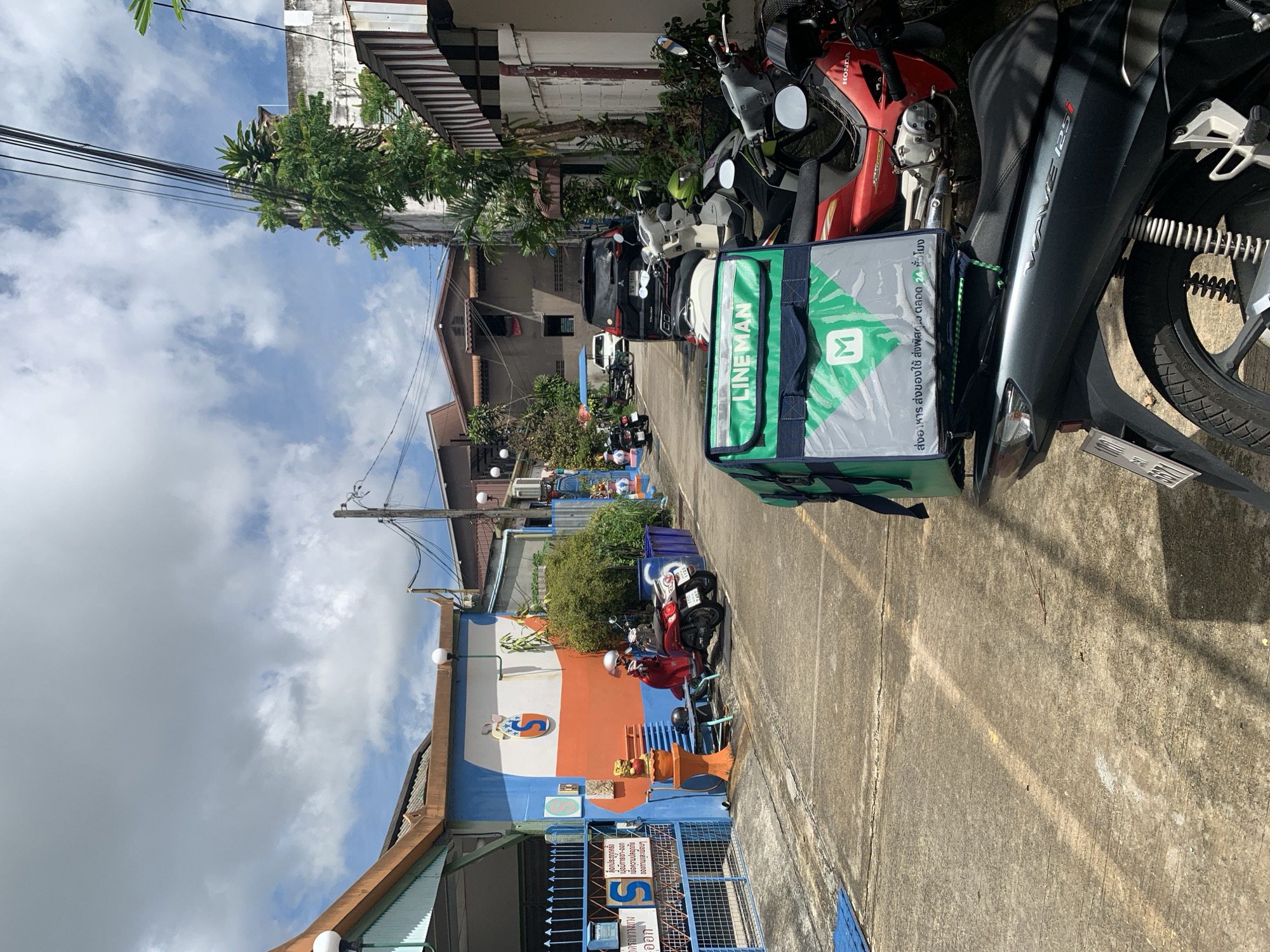 In a narrow, paved alleyway in an urban environment, a LineMan delivery bag rests on the seat of a parked dark gray motorcycle. The bag is predominantly green and silver with the "LineMan" logo prominently displayed. The motorcycle, a Honda Wave 125i, has a license plate with the number "5671." Several other motorcycles are parked nearby, including a red one with a rider wearing a helmet. A black SUV is parked further down the alley. The buildings lining the alley are painted in bright colors, with a blue and orange facade on the left featuring a circular logo and a child's drawing. Lush green foliage is visible, indicating a possibly tropical location. The sky is partly cloudy.