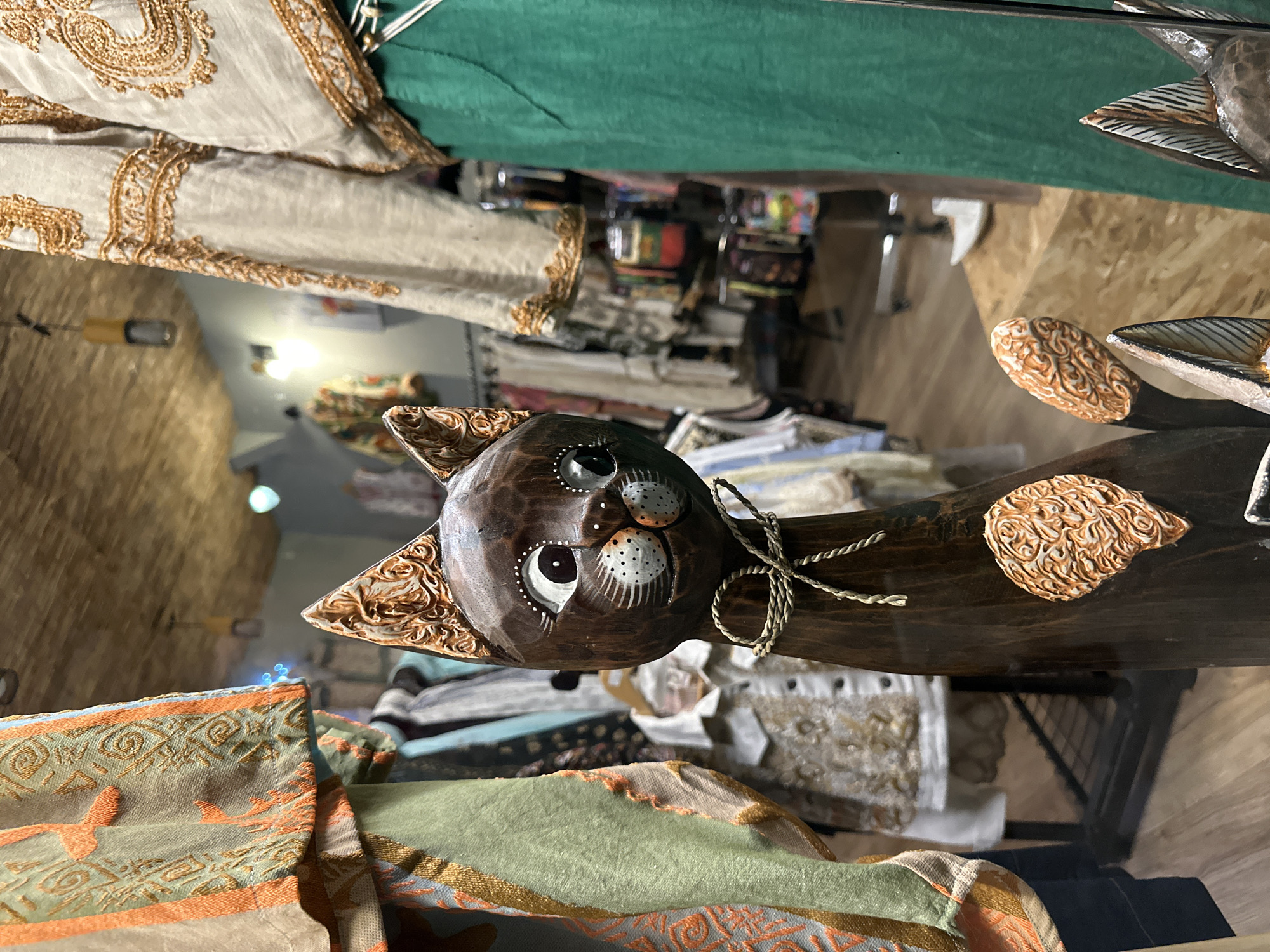 The image depicts a wooden cat figurine on display in a shop window in Veliko Tarnovo, Bulgaria. The cat is brown with carved details on its head and tail. It has large, painted eyes with white dots. A thin rope is tied around its neck. The background shows a variety of clothing and other merchandise for sale in the store. The shop window reflects the surroundings, creating a sense of depth. The scene is well-lit.