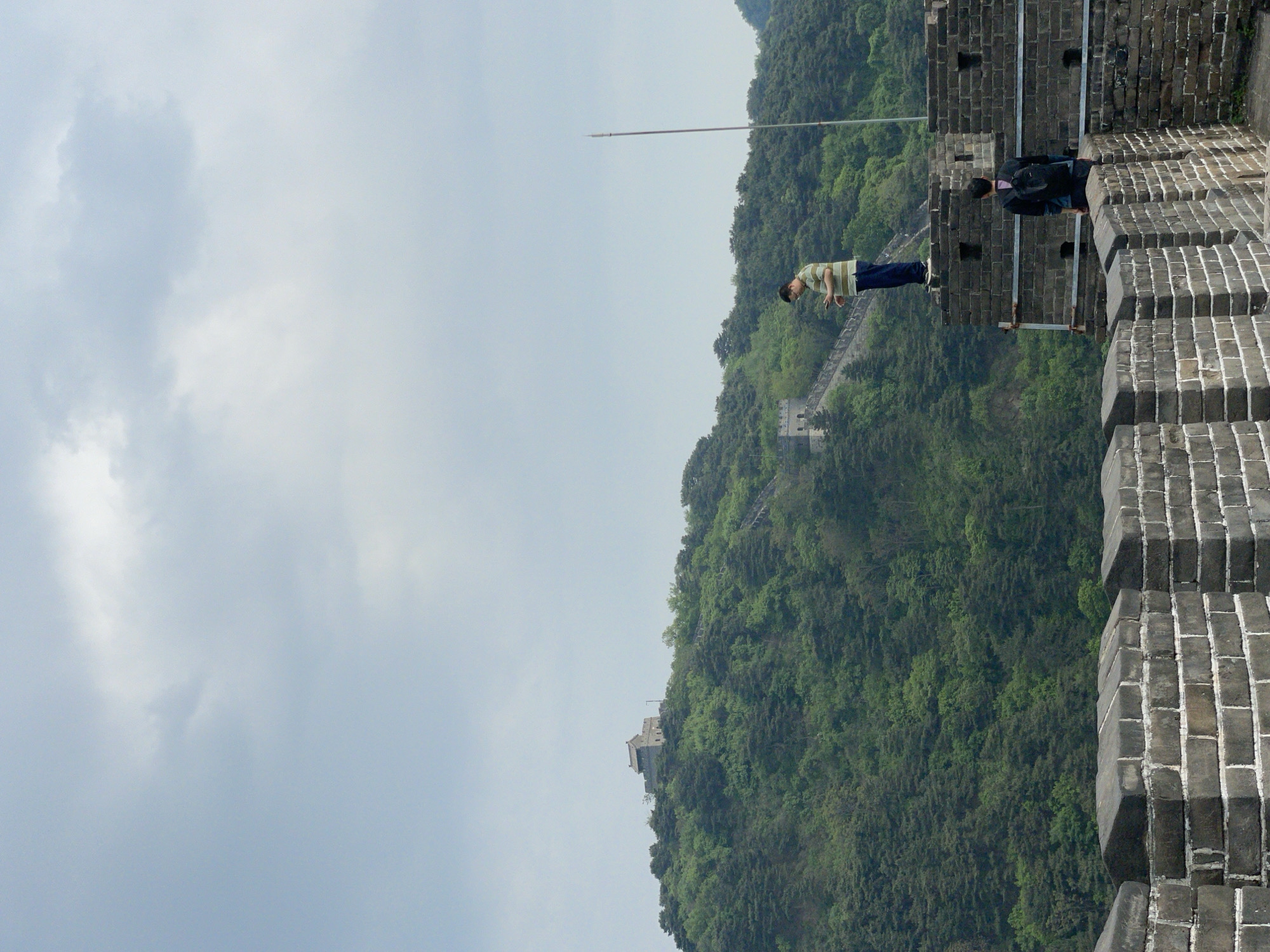 A wide-angle shot captures a section of the Great Wall of China in the Huairou District. The ancient grey brick wall, with a few people walking on it, is juxtaposed against a backdrop of a mountain covered in dense green trees. The sky is overcast. A flagpole extends towards the sky from the wall. A smaller segment of the wall is visible further down the mountain.