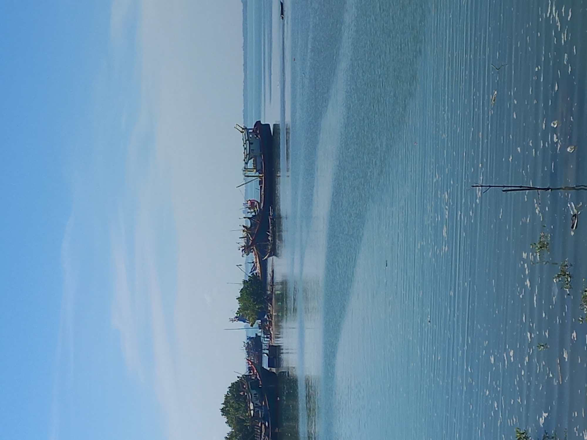 The image captures a waterfront scene in Lhokseumawe, Indonesia. Several boats are moored along the coast, adjacent to a shoreline with trees and vegetation. The water reflects the sky, which has clear patches interspersed with some cloud cover. The perspective is likely a low angle, capturing the boats and water prominently.