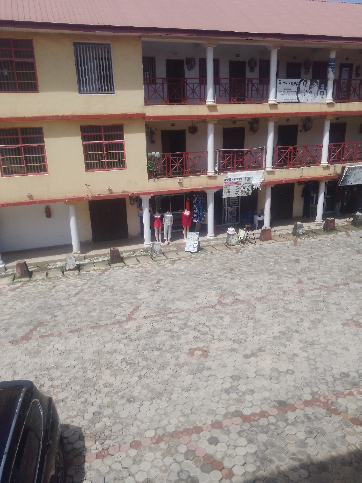 The image depicts a two-story building with a red corrugated metal roof, situated in Gura Top, Nigeria. The building has a cream-colored facade and is adorned with red trim around the windows and balconies. The ground floor features an open-air walkway with white pillars and red railings, housing several small shops. In front of these shops, mannequins are displayed, some wearing red dresses and others white attire. A black car is partially visible in the lower left corner of the image. The ground in front of the building is paved with hexagonal interlocking concrete blocks, some of which are highlighted with red accents. A sign for "IVE-LYN" is visible above one of the shops, and another sign with an image of people and text is mounted on the upper floor's balcony. The scene suggests a commercial area with retail establishments.