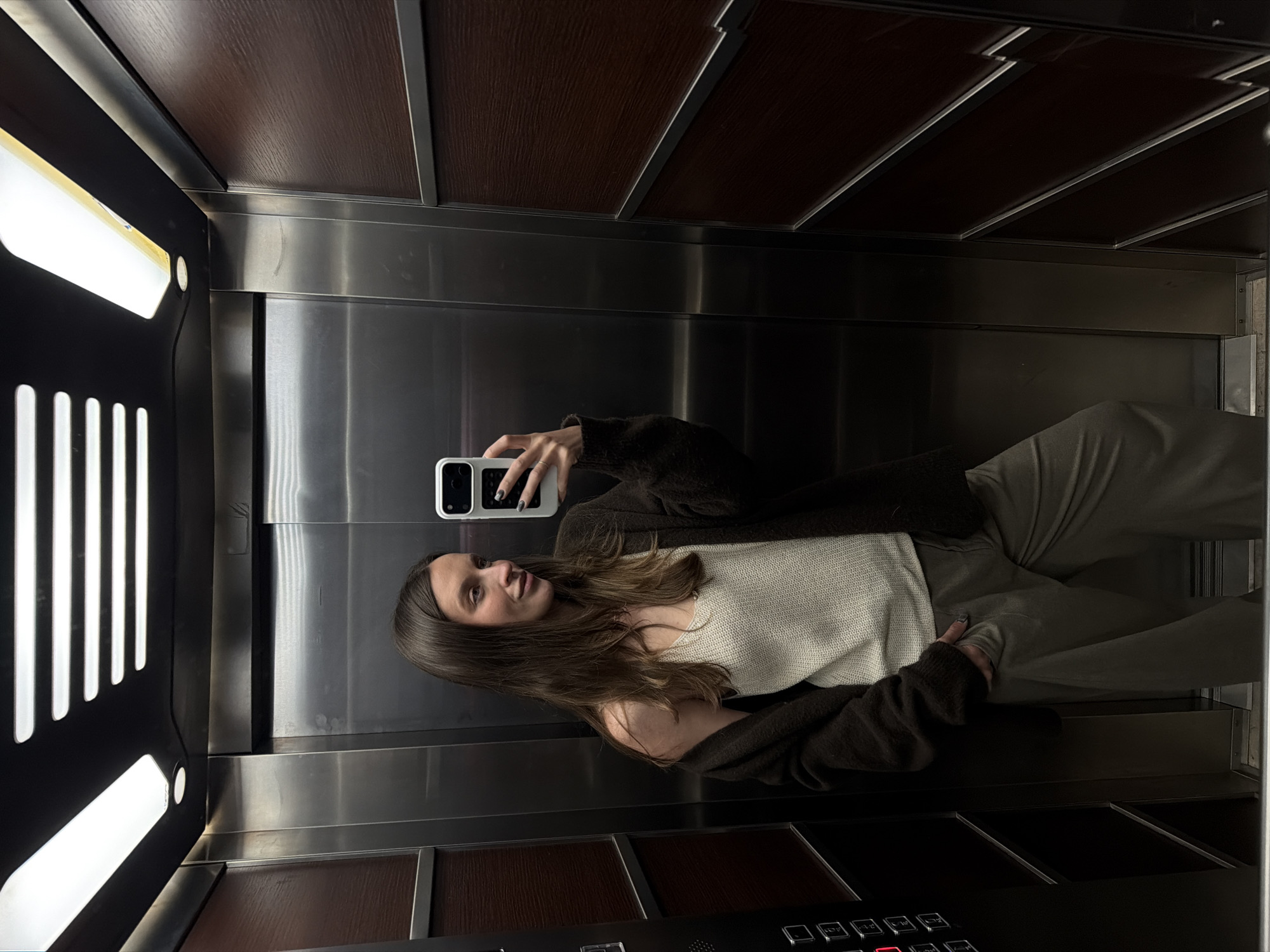 A woman is taking a selfie in an elevator. The elevator has a dark brown ceiling with metal trim, stainless steel walls, and a black control panel with buttons. The lighting is provided by a fluorescent light fixture. The woman is wearing a light-colored top, olive green pants, and a dark brown cardigan. She is holding a white smartphone in her left hand. The location of the image is unknown.