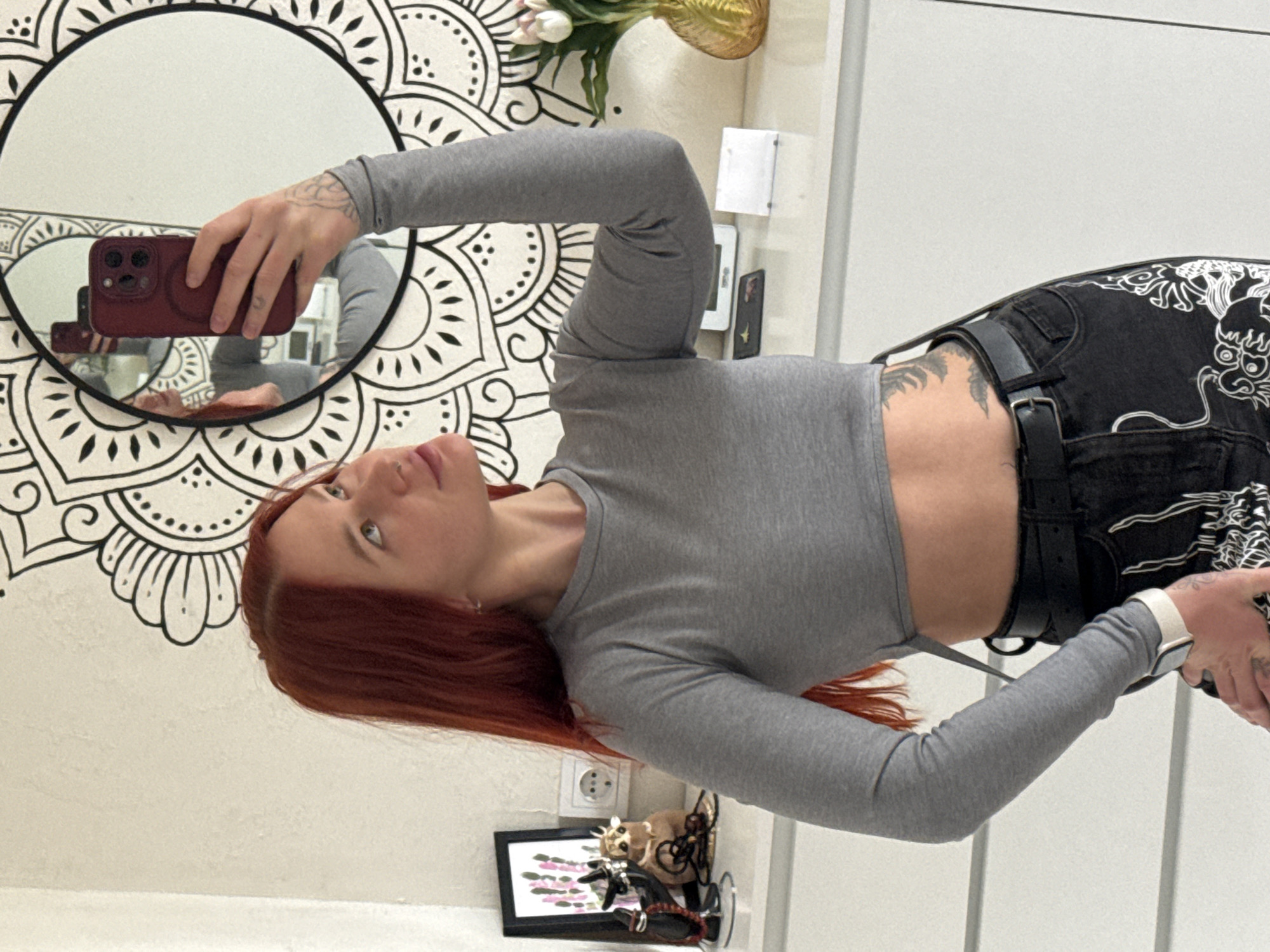 A young, fair-skinned woman with red hair is taking a selfie. She is wearing a gray long-sleeved crop top and black pants with white patterns. Two black belts with silver buckles cinch her waist. She is holding a maroon-colored phone with her left hand, visible in a round mirror with a decorative black and white mandala pattern on the wall behind it. The mirror reflects her face and the phone she is holding. Her right hand is partially visible in the lower right corner, showing a white smartwatch. Tattoos are visible on her left forearm and right hip.
Behind her, near the bottom of the frame, is a black picture frame with what appears to be a small painting of pink flowers, and a black sculpture of a cat and a dog. A European-style electrical outlet is visible on the wall next to the picture. There is also a small vase with artificial flowers and a small electronic device mounted on the wall. The wall behind her appears to be white.
The scene is indoors, possibly in a home or apartment. There is no information in the picture to indicate that this is located in Rostov-on-Don, Russia.