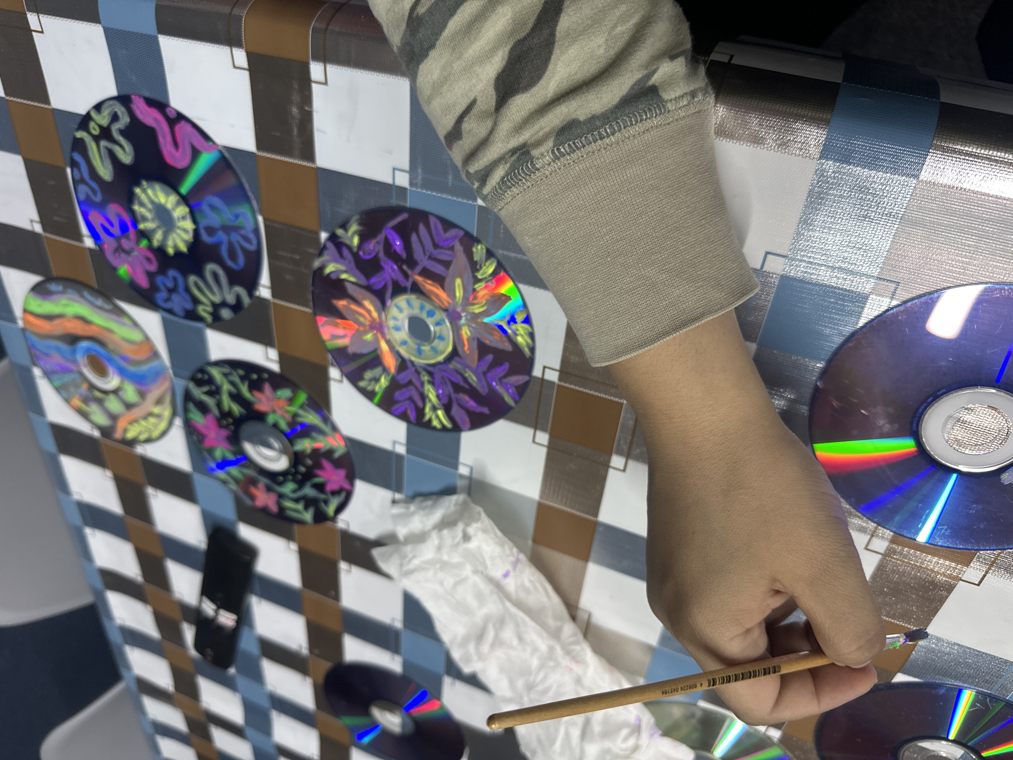 A person is sitting at a table painting designs on CDs. The table has a checkered pattern. There are several CDs with different designs on the table. The person is wearing a camouflage-patterned sleeve. A tissue is visible on the table. A pencil is held in the person's hand. There is a black smartphone on the table. The location is unknown.