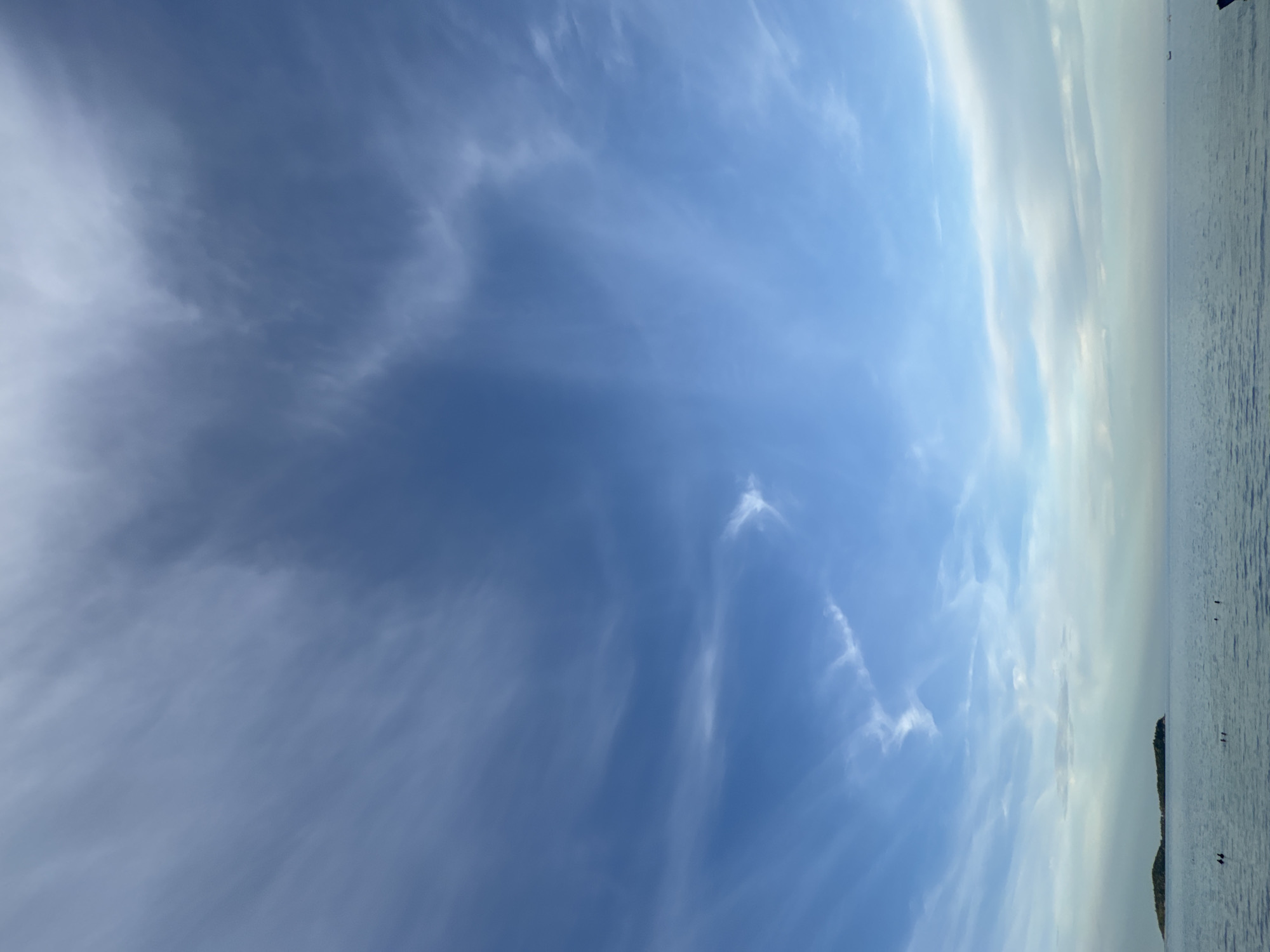 A tranquil scene unfolds where a vast expanse of calm, blue water meets a sky painted with wispy, white clouds against a bright blue backdrop. A small, green island, possibly a distant landmass or a group of islands, is visible on the left side of the horizon. The water's surface is gently rippled, reflecting the soft light from the sky. Several dark, indistinct shapes, possibly buoys or small boats, are scattered across the water. The overall impression is one of peacefulness and natural beauty.