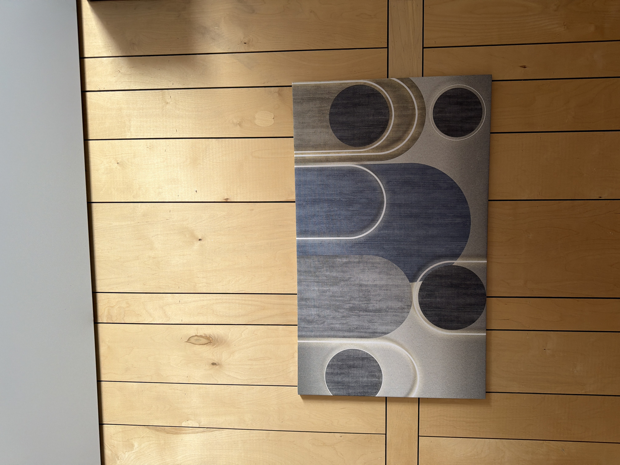 A rectangular artwork featuring abstract geometric shapes is mounted on a wall clad in vertical, light-colored wood panels. The artwork displays a pattern of circles and curved lines in shades of gray, blue, and tan. The wood paneling has visible grain and knots. Above the wood paneling is a horizontal band of a smooth, light gray surface. The lighting suggests natural light is casting shadows on the right side of the wood paneling. There are no people or animals present in the image. The scene appears to be an interior wall.