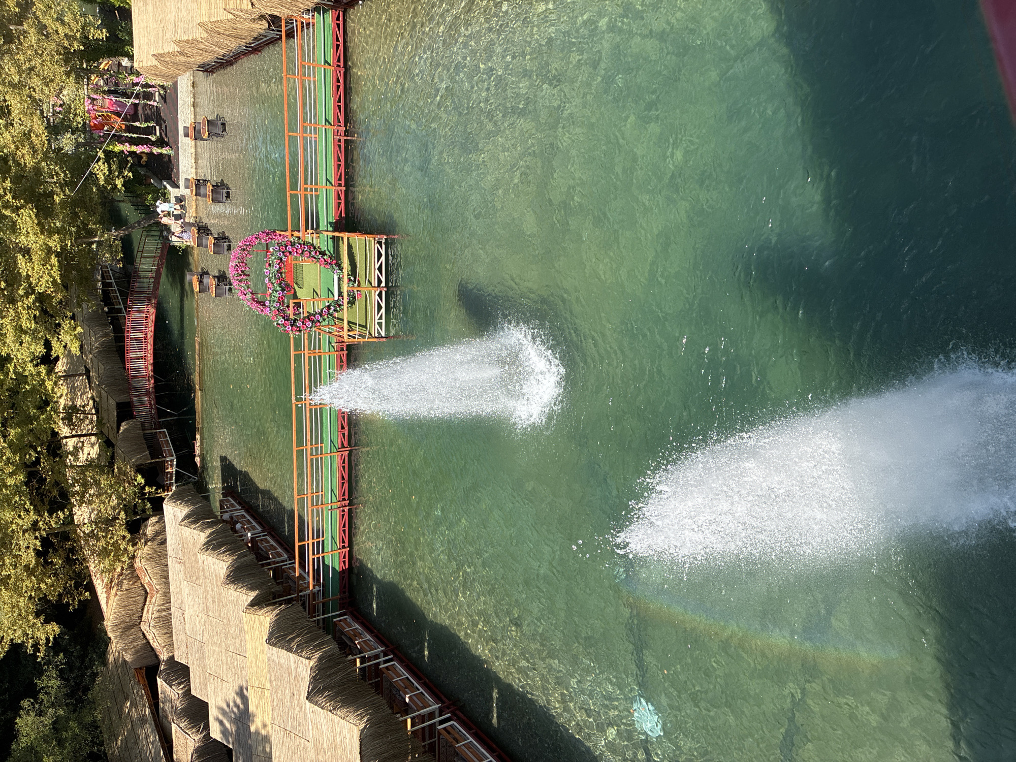 The image shows a waterfront dining area in Alanya, Turkey. Several thatched-roof huts line the water's edge, with tables and chairs visible inside some of them. A wooden walkway with red railings extends into the clear, green water. Two fountains are active in the water, creating plumes of white spray. A rainbow is visible in the mist of one of the fountains. In the background, there is a red bridge spanning a section of the water, and trees with green foliage surround the area. Some people are visible on the walkway and near the red bridge, appearing to be dining or relaxing. A heart-shaped floral decoration is positioned on the walkway between the two fountains.