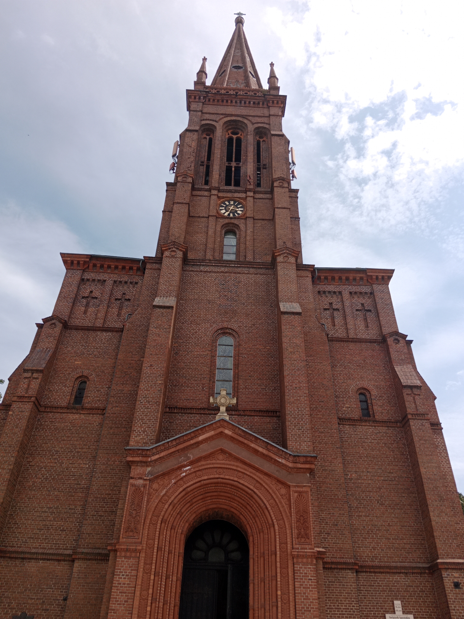 The image shows the facade of a red brick church, the Church of St. Andrew Bobola, located in Bydgoszcz, Poland. The church features a tall, pointed spire, a clock on its tower, and arched windows. The entrance to the church is a large, ornate arched doorway with decorative carvings. The building is constructed of red brick, and the sky above is partly cloudy.