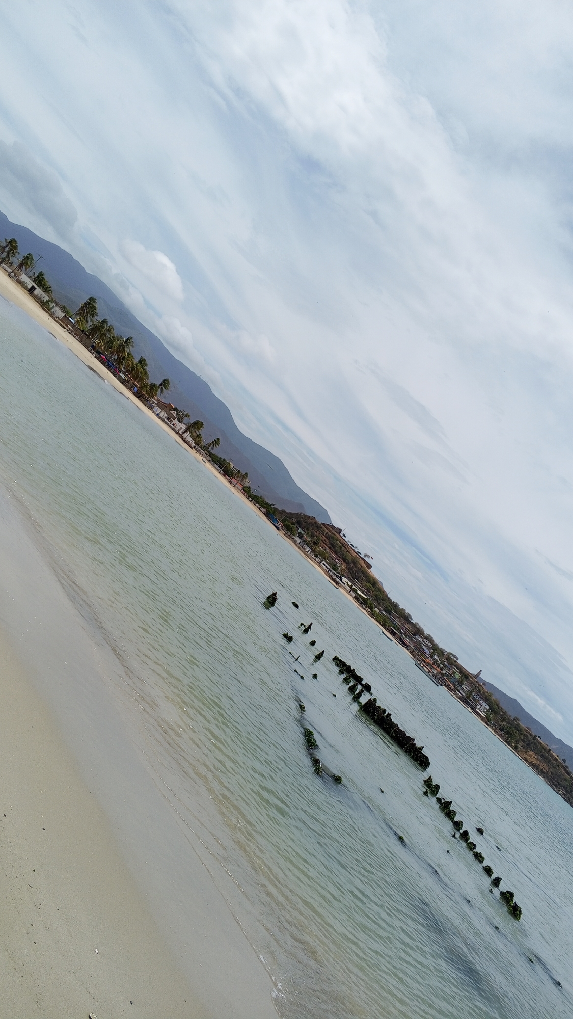 The image depicts a beach scene in Juan Griego, Venezuela, with the camera tilted at an angle.
**Environment:** The scene consists of a sandy beach merging into shallow, calm water. A line of dark posts or wooden structures, likely remnants of an old pier or breakwater, extends into the sea. These structures are partially submerged and appear to be covered with green algae or marine growth. The background includes a coastline with buildings, trees, and mountainous terrain under a sky filled with patchy clouds.
**Objects:** Key objects include the beach, water, wooden posts, buildings, trees, and mountains.
**Actions/Interactions:** The water gently laps onto the beach. The scene suggests a tranquil coastal environment.
**Location:** Juan Griego, Venezuela. The presence of the beach, mountains, and specific architectural elements of the coastline is consistent with the geography of the area.