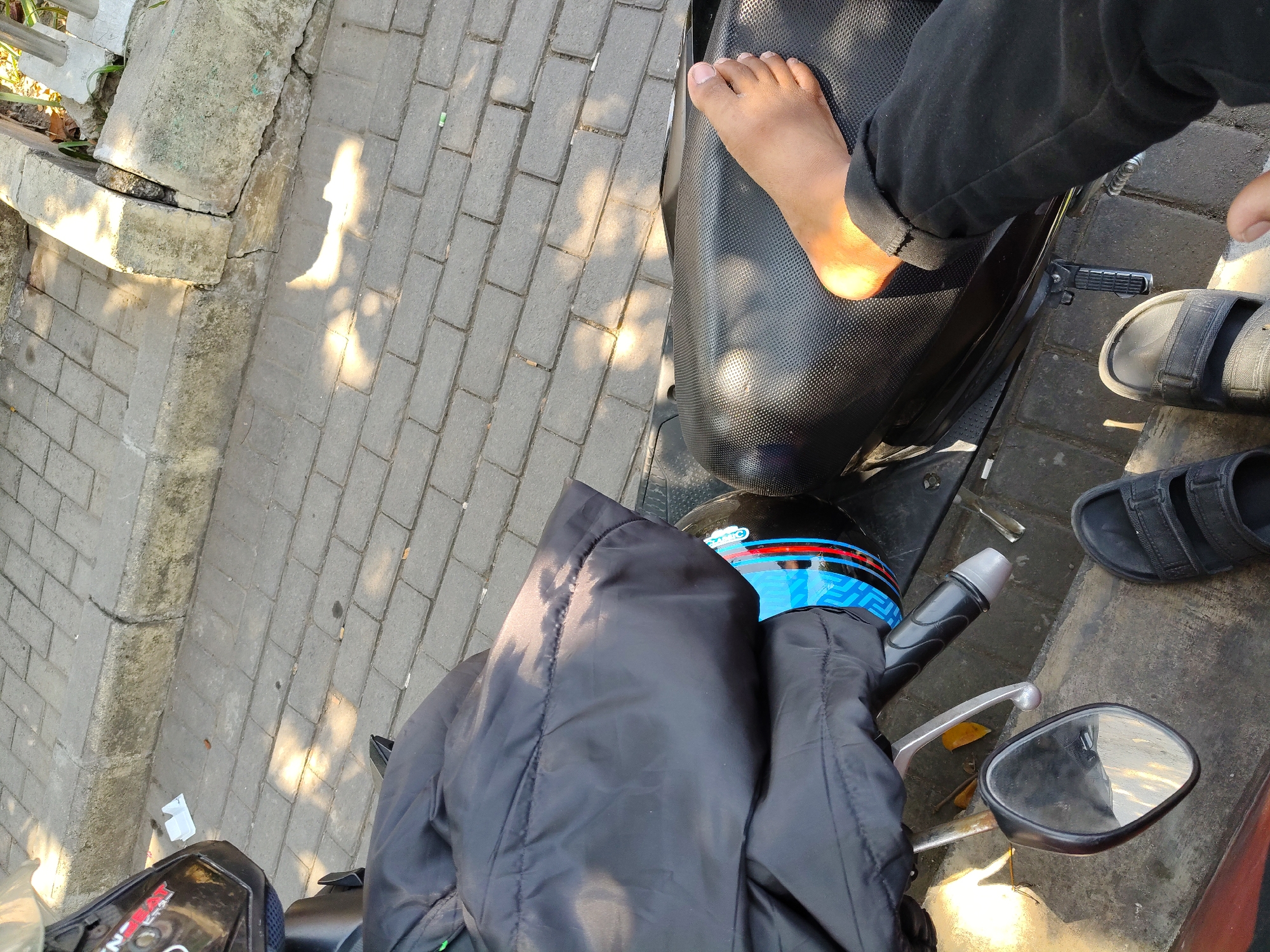 The image shows a person's feet in black sandals and black pants, with a bare foot and leg on a black motorcycle seat. A black jacket is draped over the motorcycle. A black helmet with blue and red stripes is visible. The motorcycle is parked on a brick sidewalk next to a concrete curb. The environment appears to be an outdoor urban setting, possibly in Surabaya, Indonesia, given the context. Sunlight casts shadows on the brick pavement, indicating a bright, sunny day.
