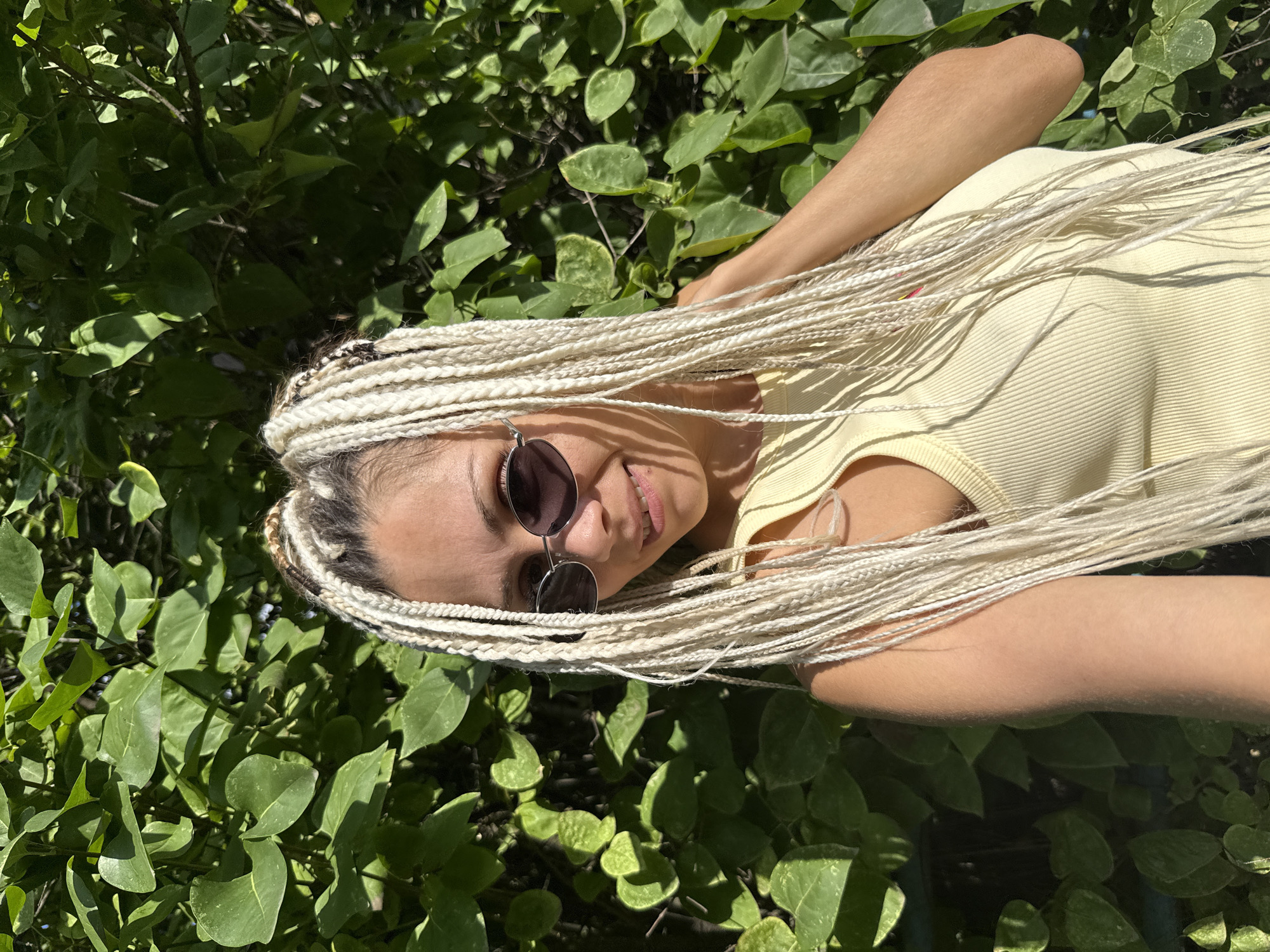 A woman with blonde braids and sunglasses is standing in front of a lush green bush. She is wearing a yellow tank top and is smiling at the camera. The sun is shining, casting shadows on her face and the bush. The location is not specified as Rusanovo, Russia.