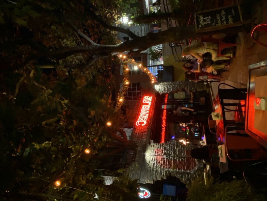 A nighttime scene depicts an outdoor restaurant area. The red neon sign "CERVELAR" is prominently displayed on a stone wall, illuminated by string lights. Patrons are seated at tables, with some appearing to be enjoying their meals. Vegetation, including trees and bushes, frames the scene. A menu board advertising "2x1" specials is visible. The overall ambiance is warm and inviting.