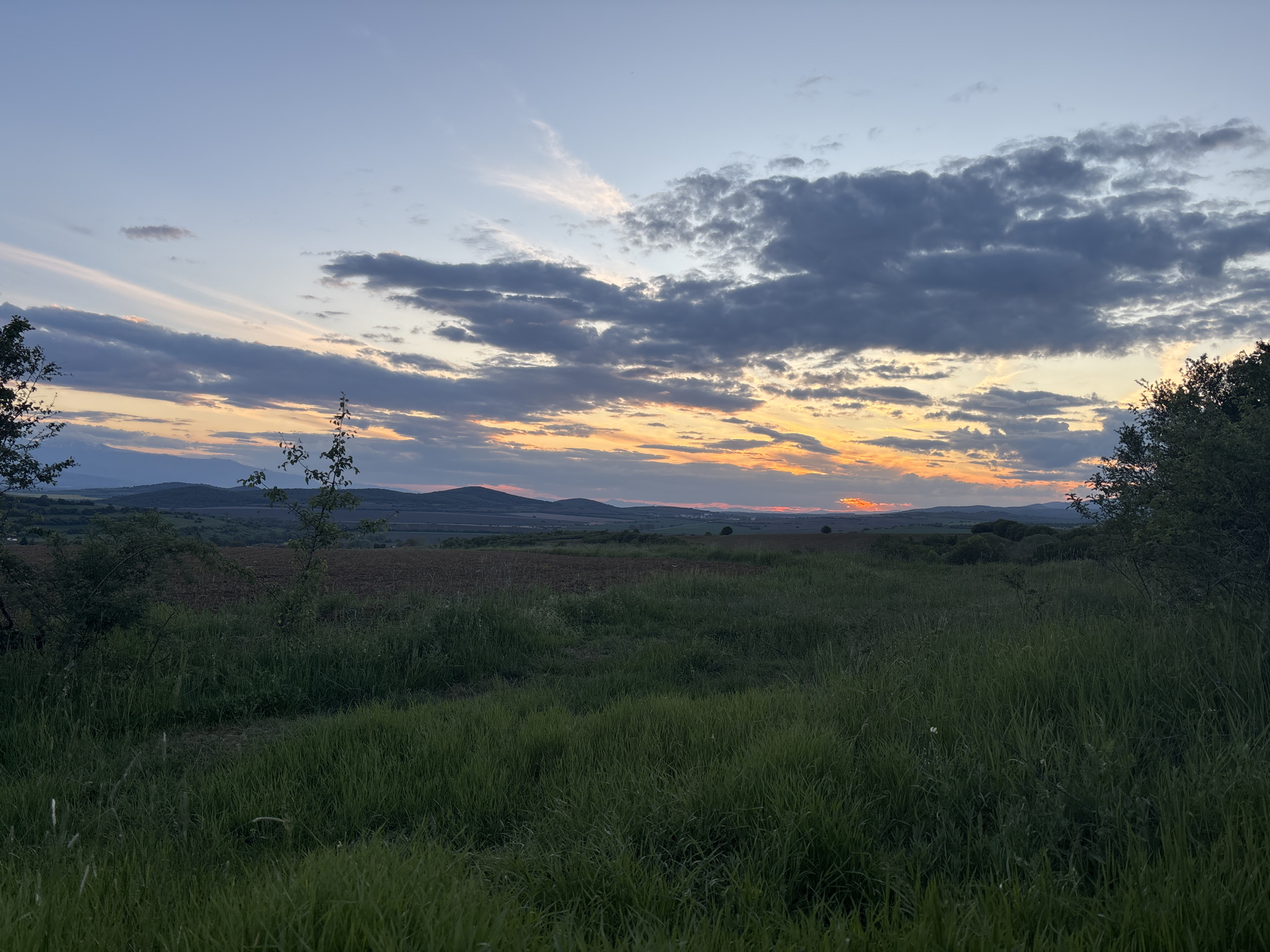 The image shows a landscape scene taken near Belopoptsi, Bulgaria, at sunset. In the foreground, there is a field of green grass with a path or worn area visible. Beyond the field is a darker patch of ploughed land. In the midground, a line of hills is visible against the horizon. The sky above is filled with clouds, some dark and heavy, others lighter with streaks of orange and yellow hues from the setting sun. Trees are scattered around, particularly on the left and right edges of the frame. No people or animals are visible.