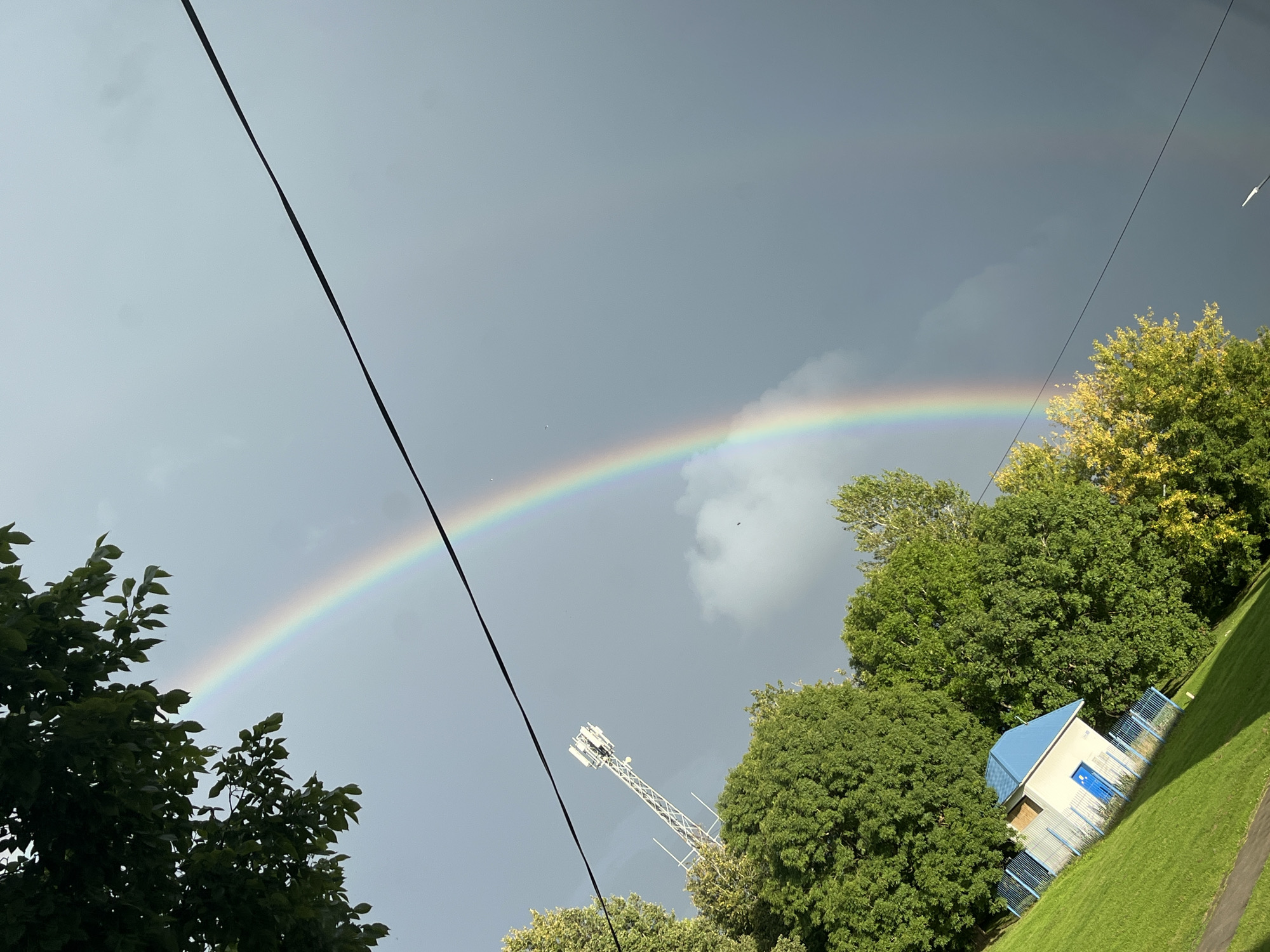 A wide, slightly tilted shot captures a sky dominated by a grey, overcast atmosphere, dramatically bisected by a vibrant double rainbow arc. The primary rainbow, intensely colored with bands of red, orange, yellow, green, blue, and violet, stretches across the frame. A fainter, secondary rainbow is visible to the right of the primary one.
In the foreground, a neatly mown green lawn slopes upwards towards a small, white building with a blue, pitched roof and a blue door. The building is surrounded by a blue metal fence. To the left of the building, lush green trees form a dense canopy, with some leaves intruding into the upper left corner of the frame. To the right and behind the building, more trees are visible, some with yellowing leaves, suggesting a transition into autumn.
In the middle ground, a tall, white lattice telecommunications tower stands prominently behind the trees. A dark electrical wire stretches diagonally across the upper portion of the image, partially obscuring the sky and the rainbow. The overall impression is of a park or outdoor public space in Kohtla-Järve, Estonia, possibly taken shortly after a rain shower. No people are visible in the image.