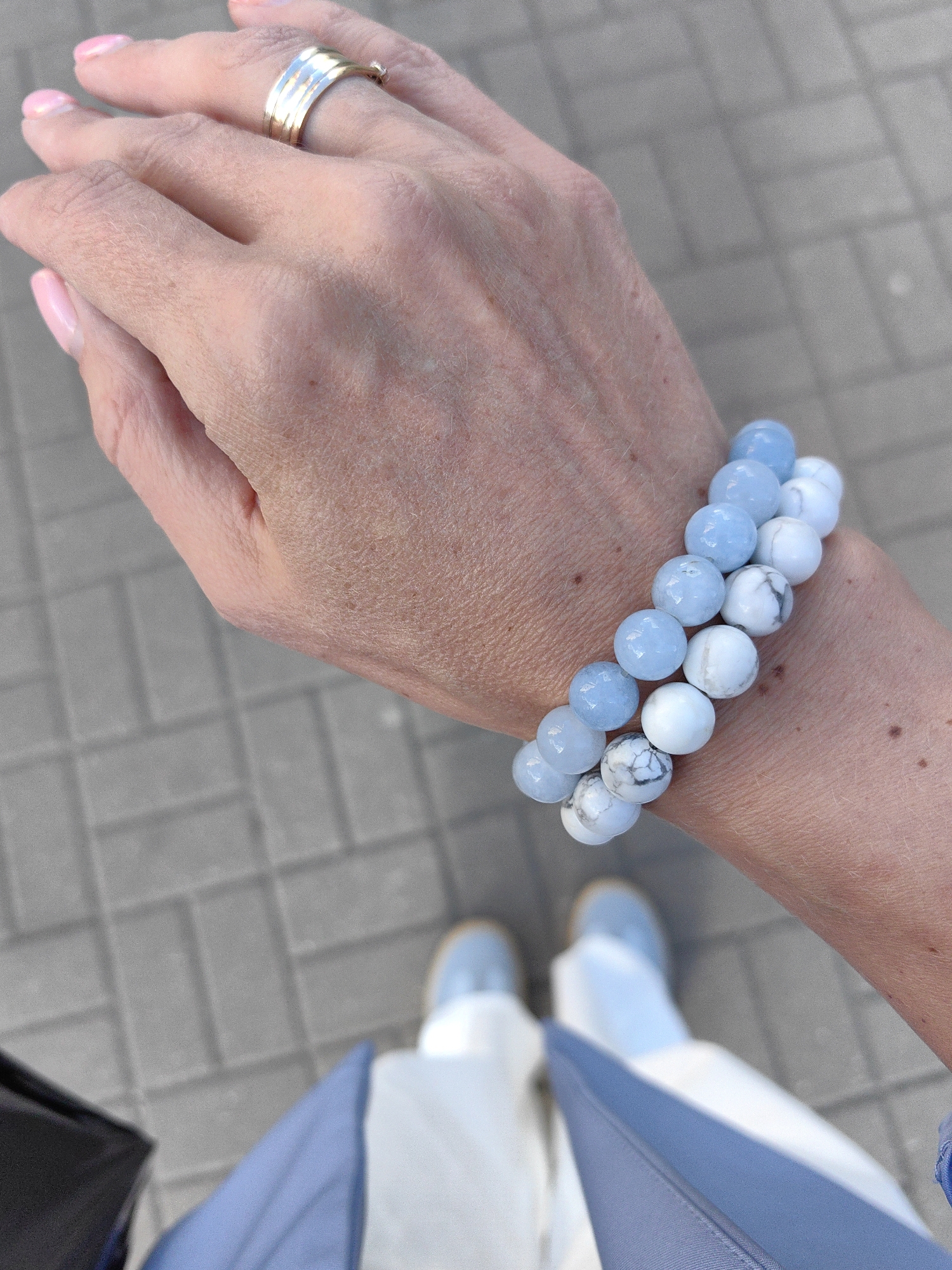 An overhead, medium shot shows a person's left hand and arm adorned with two beaded bracelets, with the lower body and feet visible in the background, all set against a paved ground. The hand, with neatly manicured pink nails, wears a simple gold band on the ring finger. The bracelets are composed of round beads in varying shades of blue and white, some with marble-like patterns. The person is wearing light blue shoes and off-white trousers, with a dark bag and blue fabric visible to the left and bottom of the frame. The scene appears to be outdoors on a paved walkway, likely in a public area.