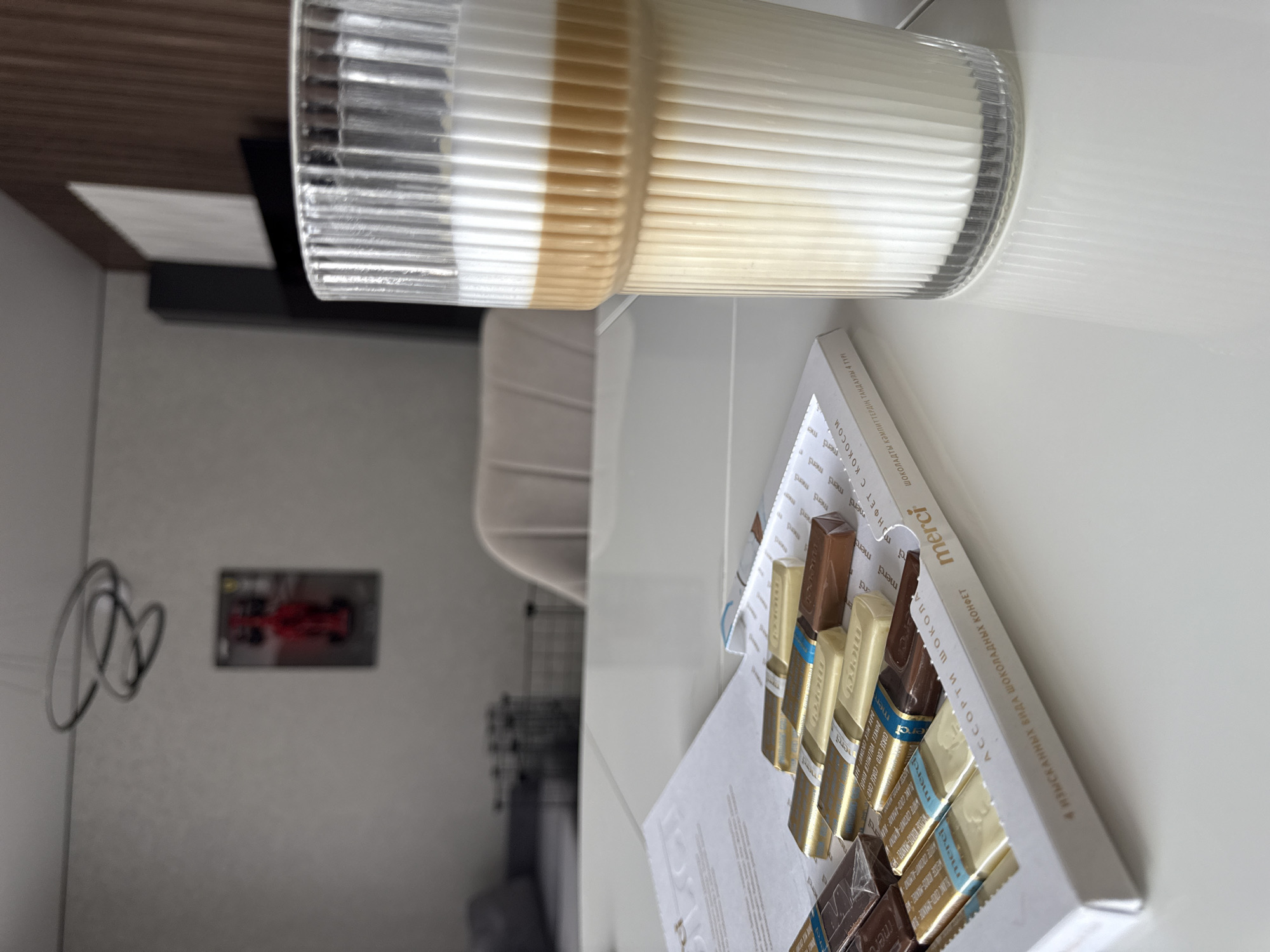An indoor scene captured from above displays a layered beverage in a clear, ribbed glass and an open box of Merci chocolates on a white surface, likely a table or countertop. The image is oriented with the items rotated 90 degrees counter-clockwise.
The tall, textured glass contains a drink with distinct layers: a lighter brown base and a thick, frothy white top layer, suggesting a latte or similar coffee drink. To the right of the glass, a white box of Merci chocolates is partially open, showcasing several individually wrapped chocolate bars in gold, brown, and blue foils.
The background is softly blurred, indicating an indoor setting, possibly a living room or office in Лобаново, Russia. Visible elements in the background include a dark wooden panel, a white rectangular object on a dark surface, and a grey wall. On the wall, a blurry rectangular image appears to depict a red racing car, possibly a Formula 1 vehicle. Nearby, some black wires form a loose loop. Further into the background, indistinct furniture shapes are visible, including what might be a chair or shelf.
The visible text on the chocolate box includes the brand "merci" and phrases in Russian: "АССОРТИ ШОКОЛАДНЫХ КОНФЕТ" (Assortment of Chocolates) and "4 ИЗЫСКАННЫХ ВИДА ШОКОЛАДНЫХ КОНФЕТ" (4 Exquisite Types of Chocolates). There is also text in Kazakh: "ШОКОЛАДТЫ КӘМПИТТЕРДІҢ ТАҢДАУЛЫ 4 ТҮРІ" (4 TYPES OF SELECTED CHOCOLATES). Labels on individual chocolates mention flavors like "WEISSE KOKOS-MANDEL" (White Coconut-Almond) and "BLANC COCO-AMANDE". The scene evokes a moment of relaxation or a casual treat.
