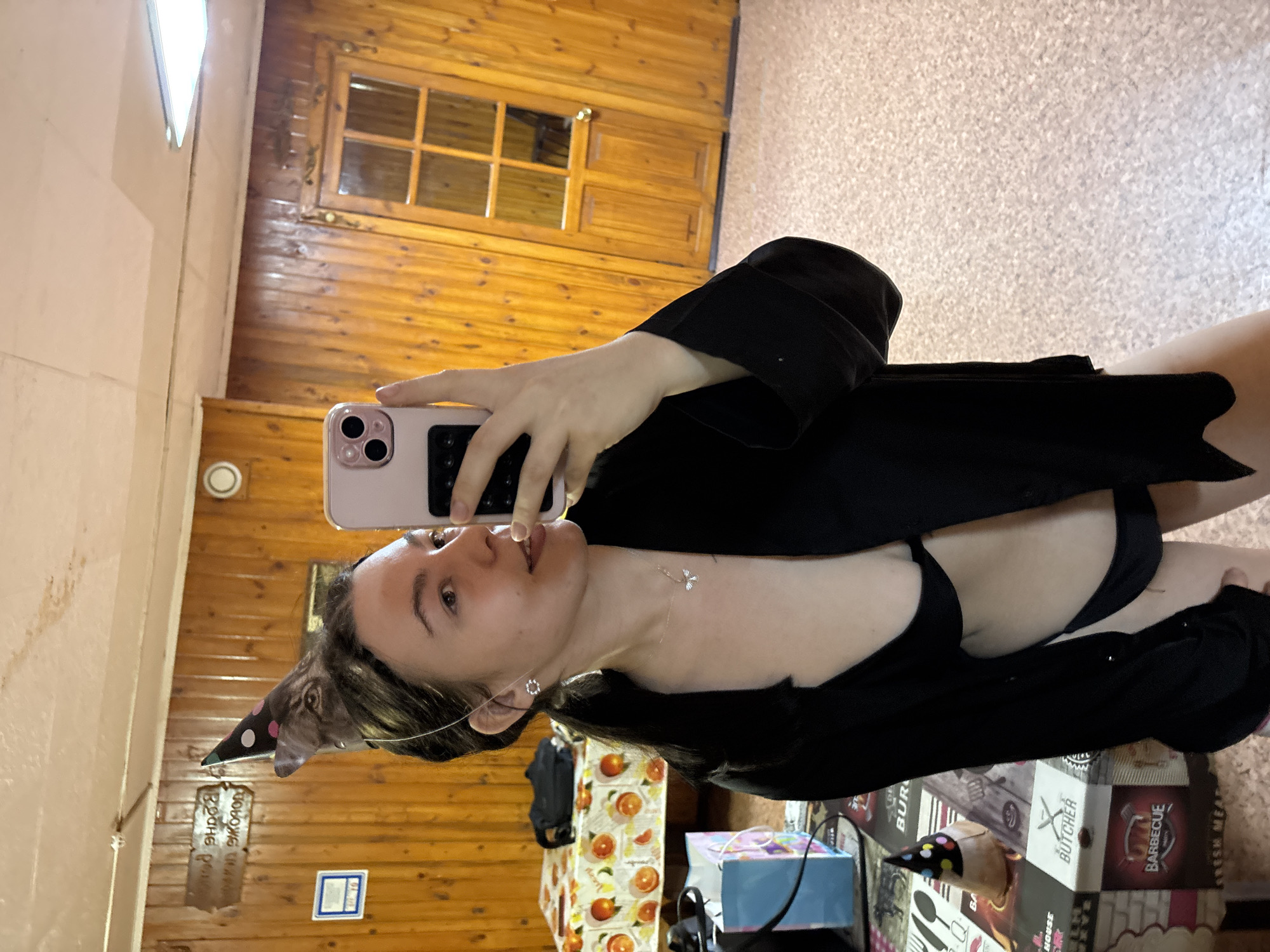 The image depicts a young woman taking a selfie in what appears to be an indoor setting. She is holding a pink smartphone with multiple lenses, partially obscuring her face. The woman has light skin, dark hair, and is wearing a black robe or cardigan over what seems to be a black bikini top. She has a necklace and earrings. On her head, a small cat is wearing a polka-dotted party hat.
The background consists of wood paneling on the walls, with a window visible in the upper portion. A portion of the floor is covered with a patterned material, possibly tile or linoleum. Near the woman, there are various items including a shopping bag with an image of oranges, a light blue box, and boxes with kitchen-related graphics, including the word "BUTCHER." There are no discernible features to definitively identify the exact location, though the prompt suggests Chaikovsky, Russia. There is nothing to independently corroborate this claim.