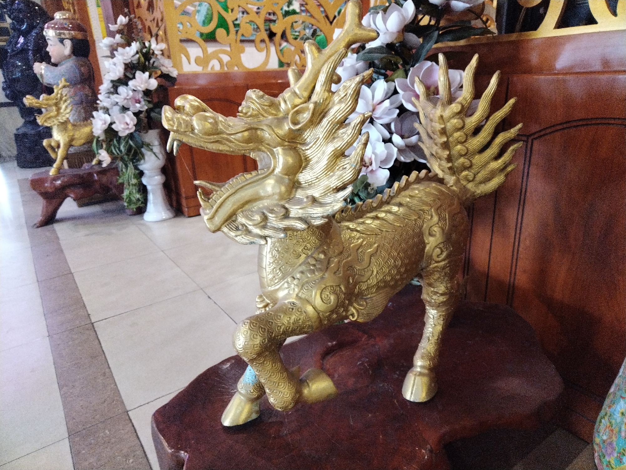 A detailed image captured indoors in Chu Văn An Ward, Vietnam, features a golden, intricately detailed sculpture of a Kirin, a mythical hooved creature, prominently positioned on a dark-stained wooden platform. The Kirin statue has a dragon-like head with an open mouth and flowing mane. The sculpture has a horse-like body covered in intricate patterns and a tail that resembles a fan of feathers.
In the background, elements such as a smaller statue of a child riding a Kirin, a white vase filled with white flowers, and a wooden screen add depth to the composition. A tiled floor in the foreground and a dark wooden panel further back provide texture and visual contrast. The lighting appears to be diffused, highlighting the details of the sculpture and the surrounding objects.