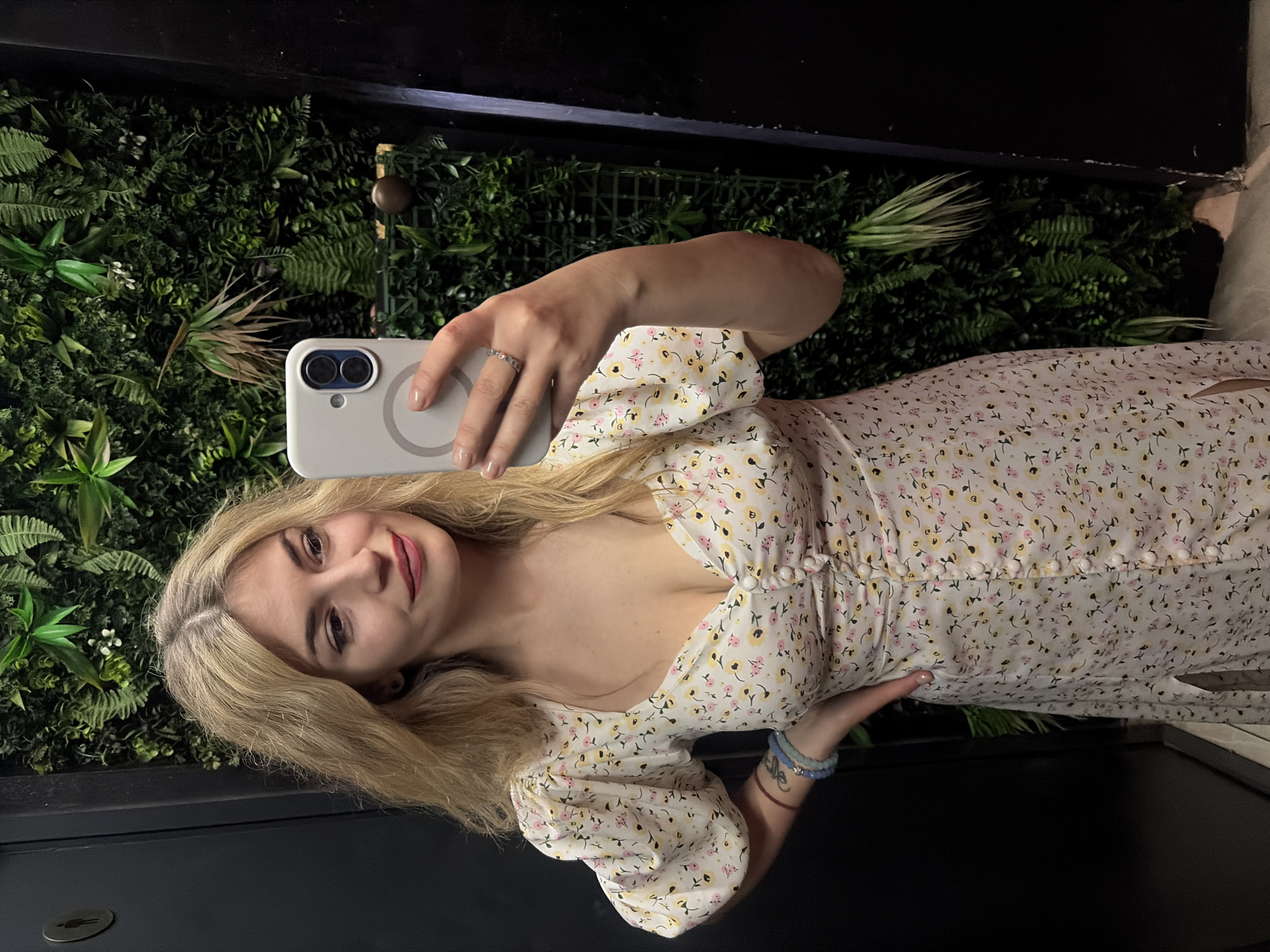A young woman is taking a selfie. She is wearing a light-colored, floral dress. She is holding a white cell phone with two lenses. She has blonde hair with darker roots. There is a green plant wall behind her. She is wearing a ring on her left hand and bracelets on her right wrist. The scene also includes a dark-colored door or wall panel. The location is Budva, Montenegro.