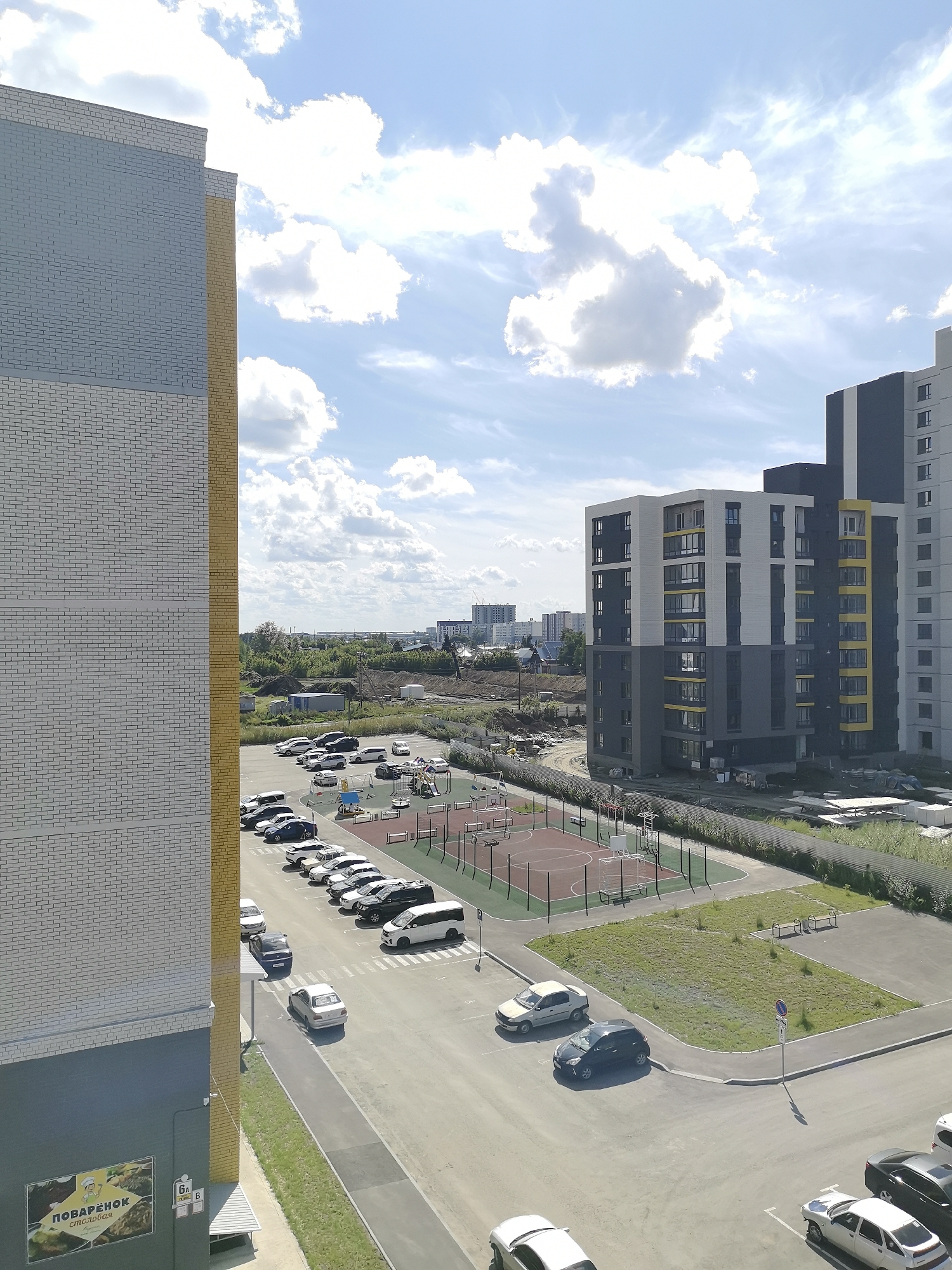 This is an aerial, medium-shot, eye-level, full-color photograph of a residential area in Novoaltaysk, Russia, on a bright, partly cloudy day. The foreground is dominated by the side of a multi-story, modern apartment building with a brick facade of light grey and yellow. To the right of this building, a complex of taller, modern apartment buildings with dark gray, white, and yellow accents is visible. Between the buildings, a paved parking lot with numerous cars is situated. Adjacent to the parking lot, a fenced-in recreational area with a basketball court, playground equipment, and benches is present. The background reveals a cityscape with more buildings, trees, and a distant horizon under a blue sky with scattered white clouds.