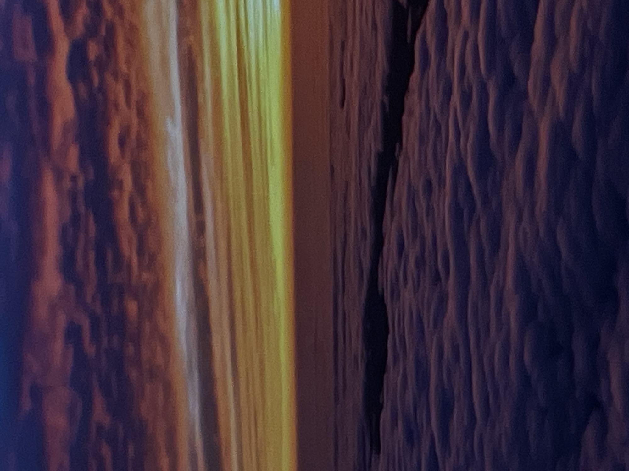 The image depicts a dramatic sunset viewed from above a thick layer of clouds. The sky is a canvas of vibrant colors, transitioning from deep purples and reds at the top, through fiery oranges and yellows, to a soft, pale yellow and greenish hue near the horizon. The clouds below are illuminated by the setting sun, creating a textured, undulating landscape of purplish-pink and dark gray shadows. The overall impression is one of ethereal beauty and vastness. There are no visible structures, people, or objects that would indicate a specific location within Denpasar, Indonesia, or any city. The scene is purely atmospheric, showcasing a natural phenomenon.